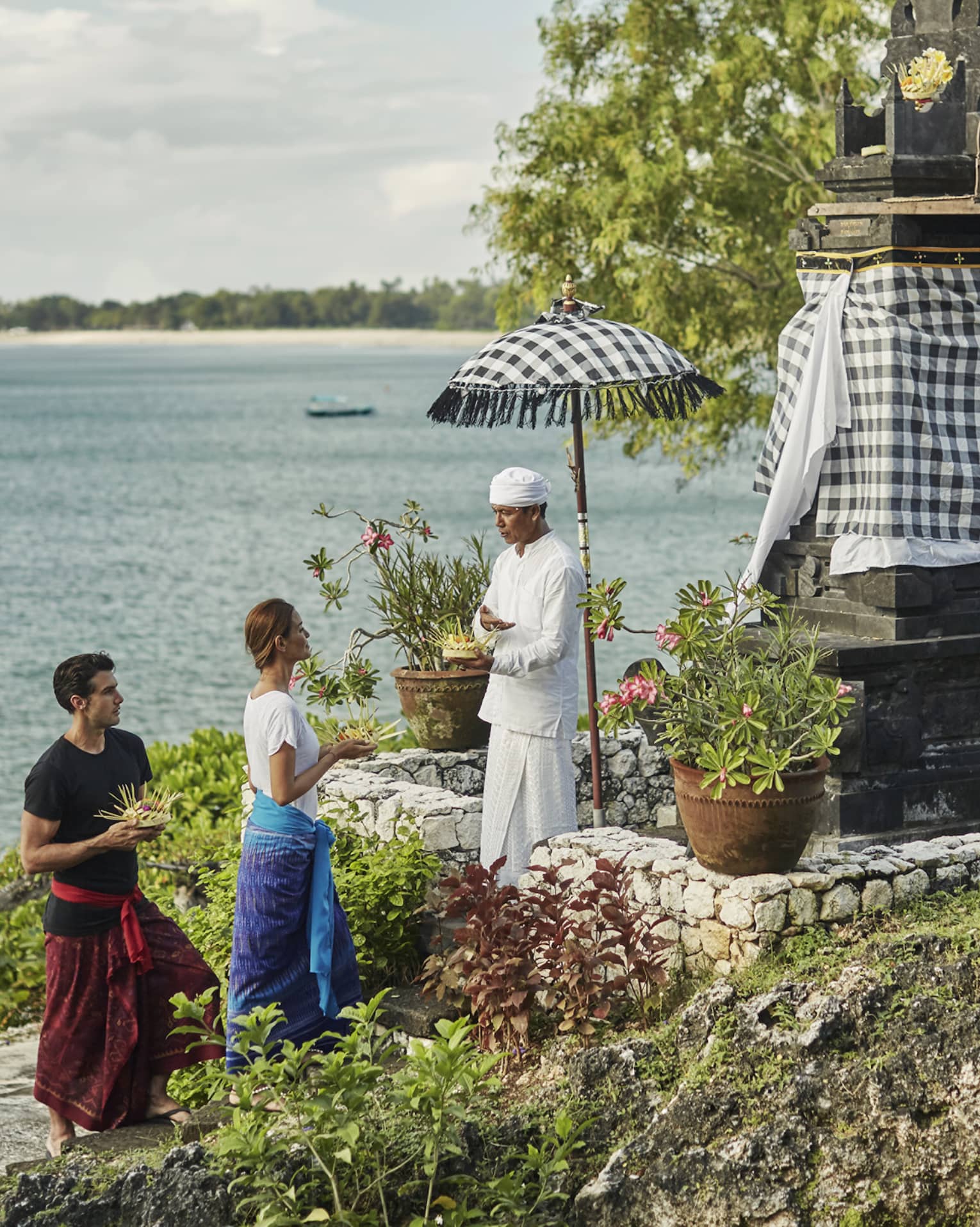 High Priest Aji Ngurah stands by shrine with black-and-white checkered umbrella, cloth, as man and woman make offering