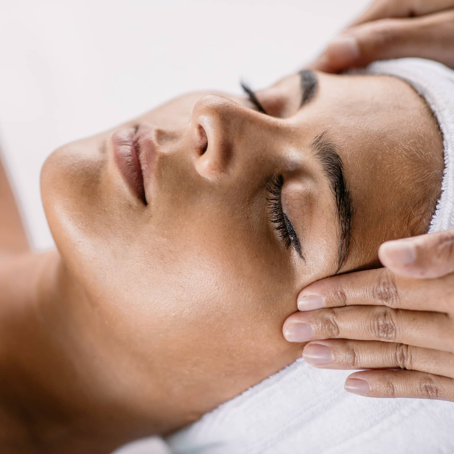 Hands massage woman's temples as she closes her eyes, spa towel around hair