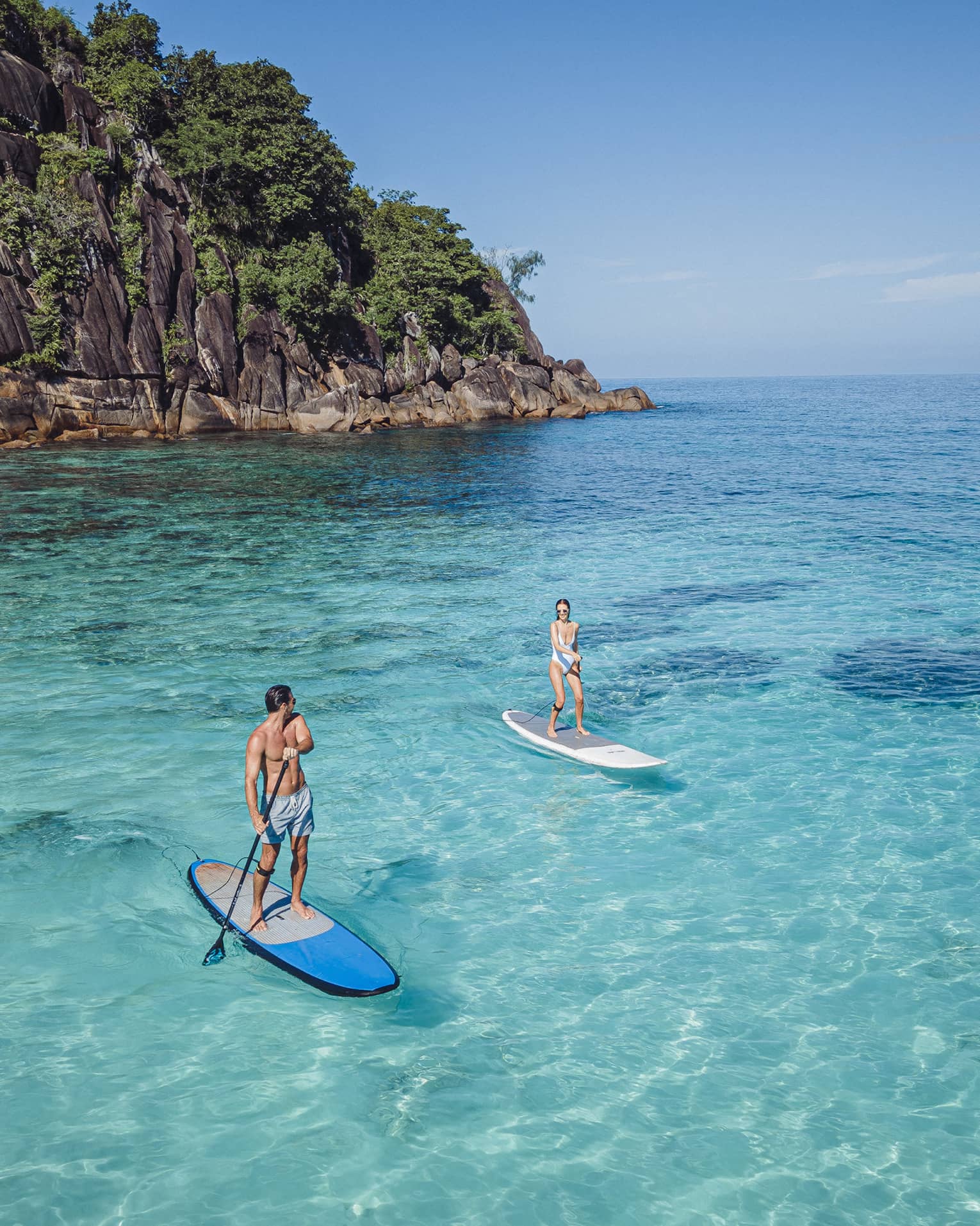 Two people stand-up paddleboarding on clear blue water with rocky cliff in back drop