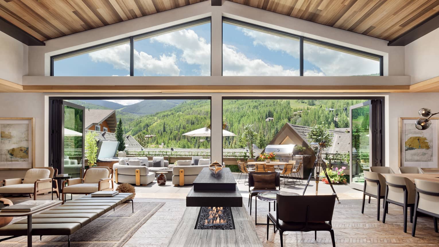Luxurious mountain villa interior with an open-plan living area, modern fireplace, and expansive glass doors leading to an outdoor terrace with panoramic forest views.