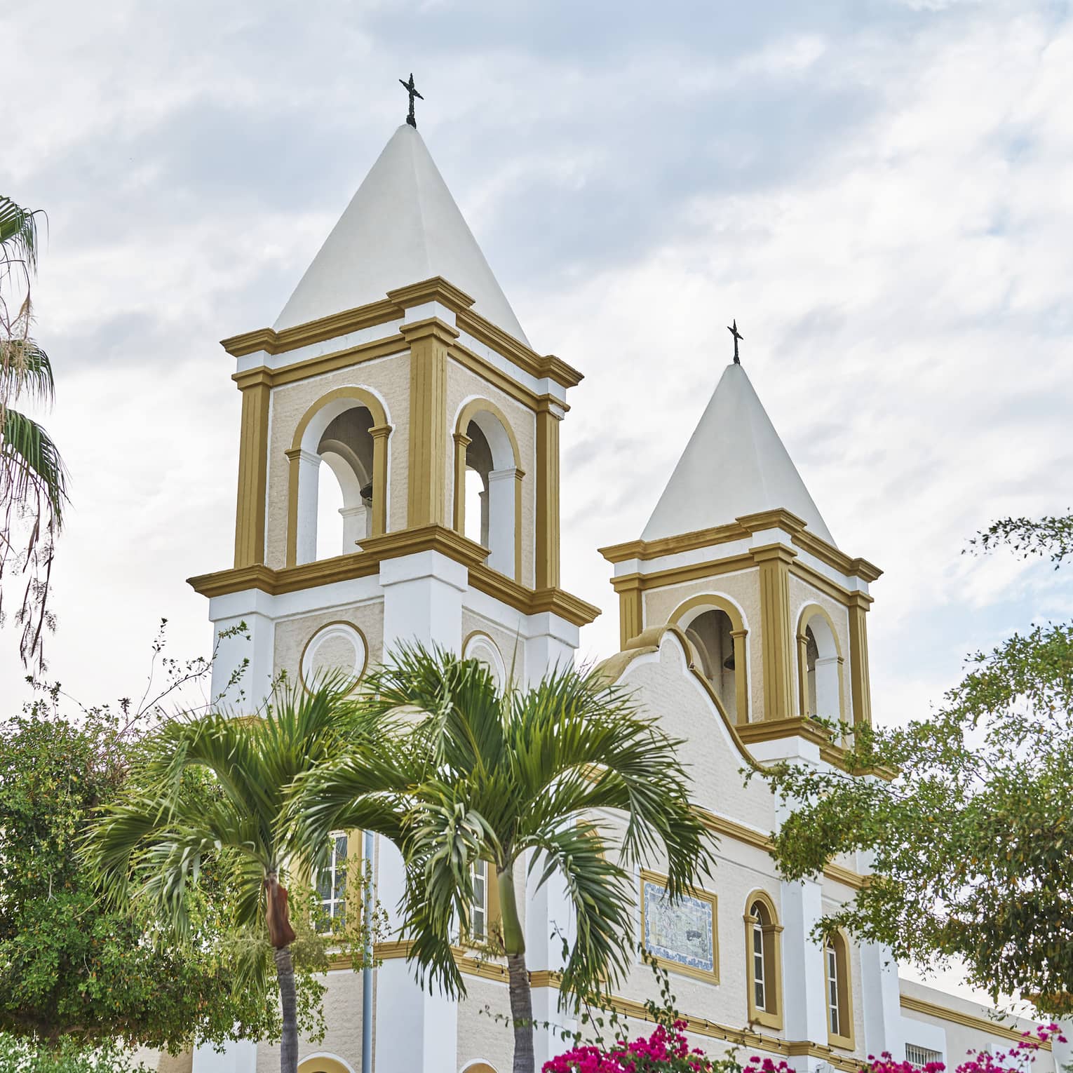 Two church bell towers rise behind palm trees and in front of a cloudy sky. They feature arched windows and gold paint.