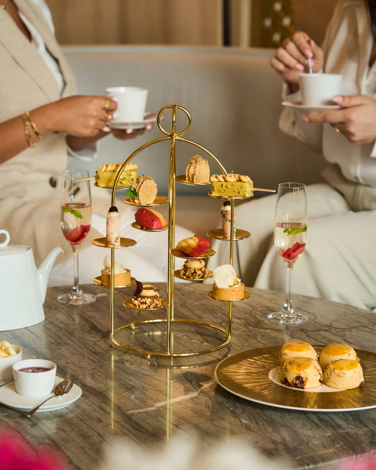 Two people sit on a beige sofa next to a marble coffee table set with a golden tray holding a variety of pastries, a white tea pot and othe small plates of food