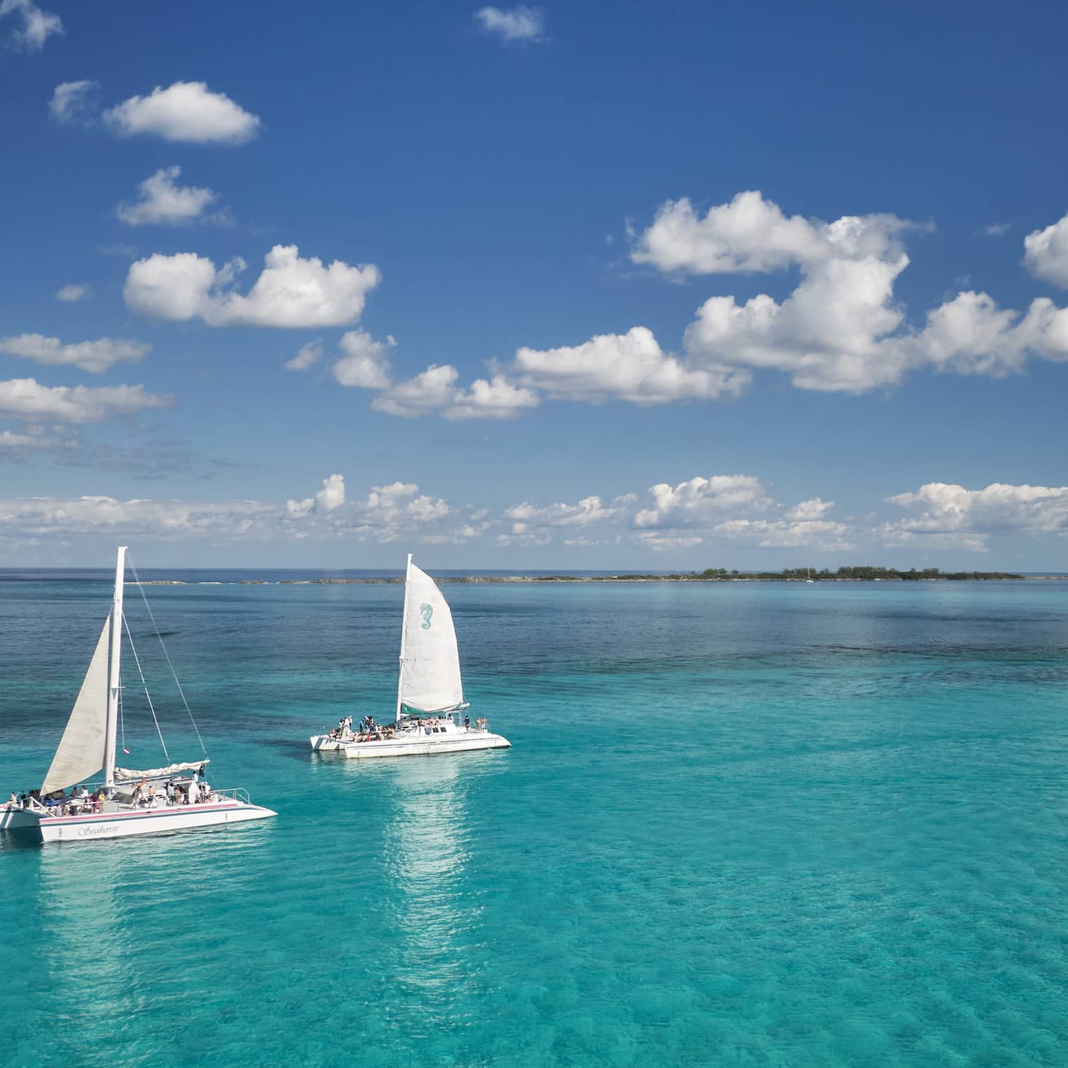 Guests aboard two catamaran sailboats glide through bright turqoise water, distant island beyond, under a cloud-dappled sky. 