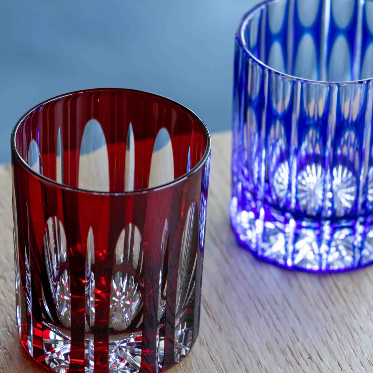 Close-up of two cut-glass tumblers with intricate geometric patterns overlaid by coloured glass, one dark red, one blue.