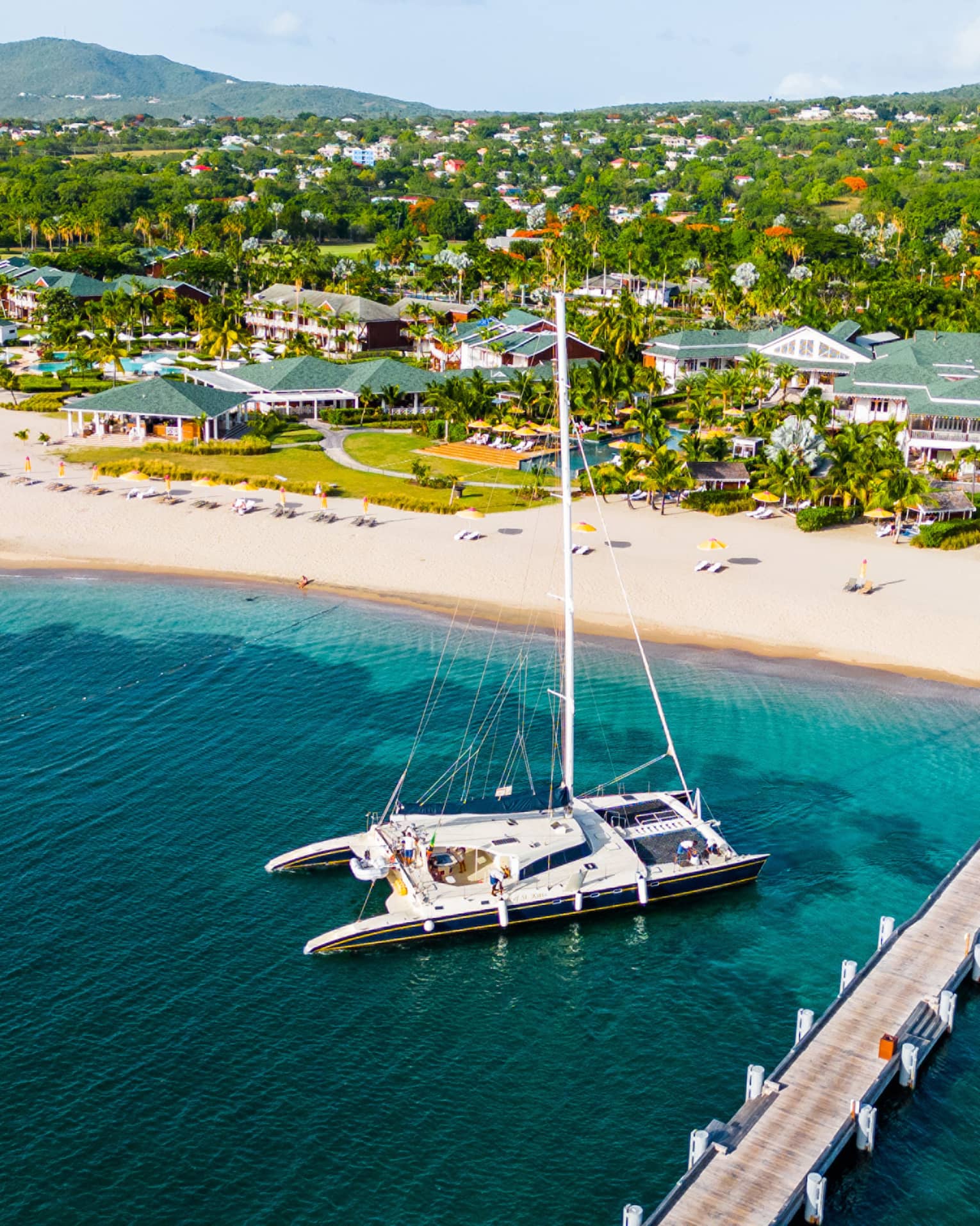 Aerial view of catamaran anchored near a dock reaching out from a beach with a sprawling resort along the coastline set among the trees