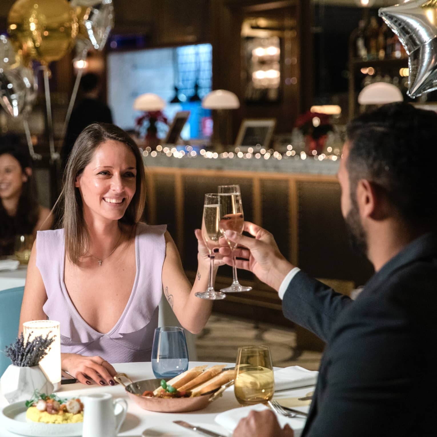 Two people clink glasses at a white linen table surrounded by balloons.