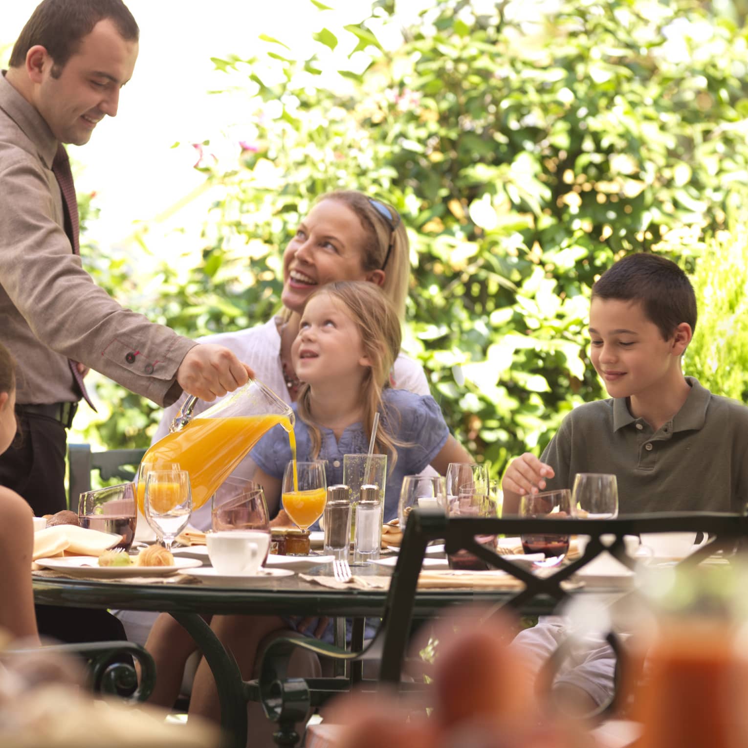 Family brunch on outdoor patio table, staff pours orange juice for young girl 