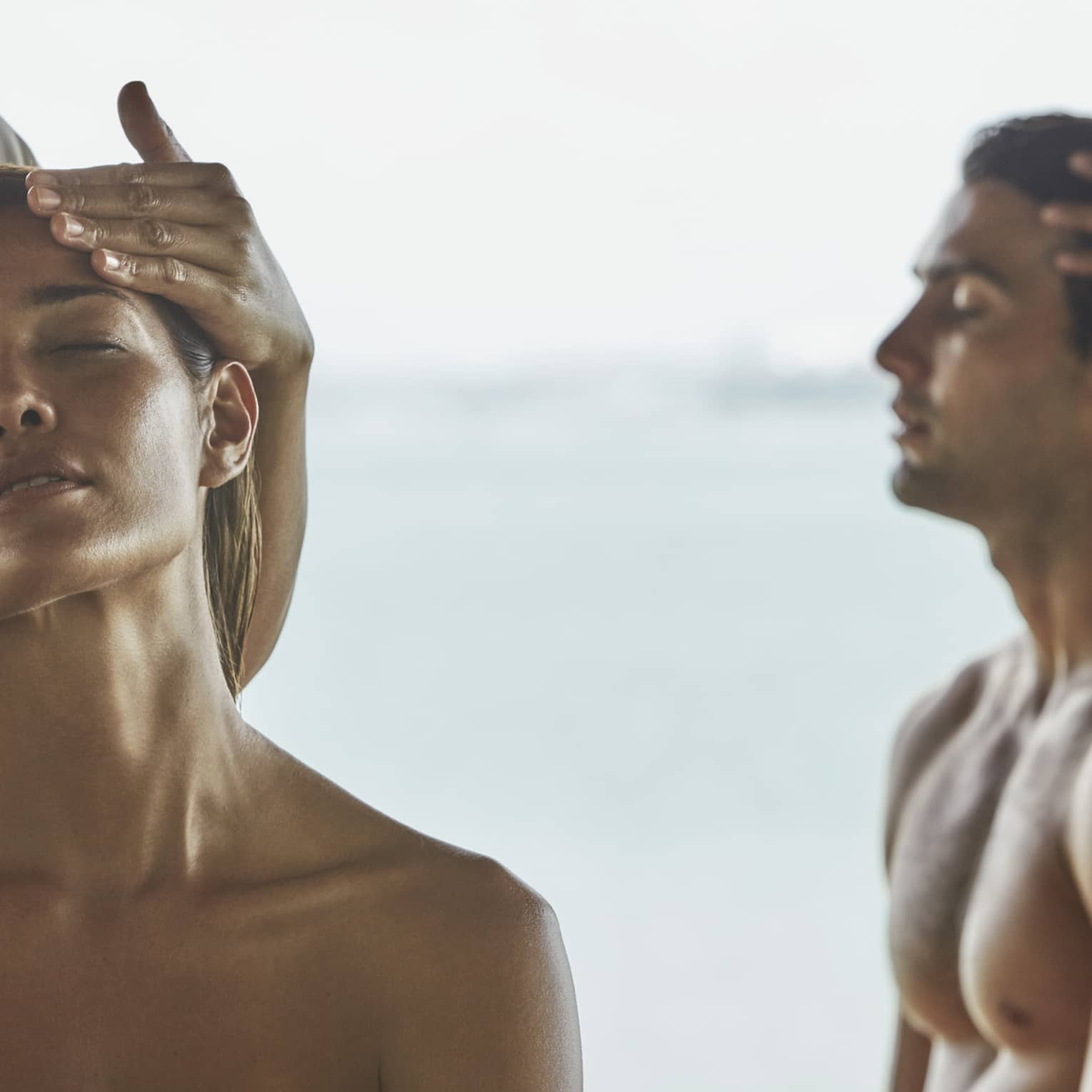 Close-up of woman and man with eyes closed as masseuses massage their temples