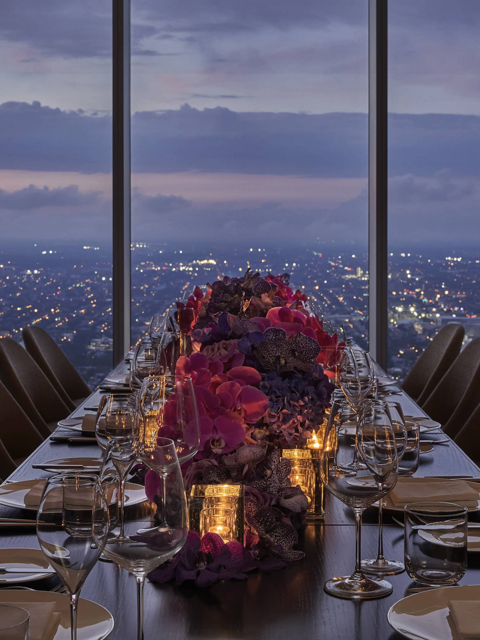A long dining table with red flowers in the center, a sun set can be seen above a city through a large window.