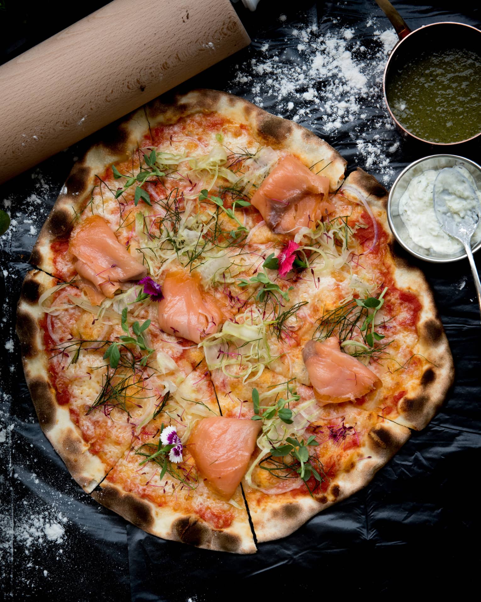 A wood-fired pizza topped with smoked salmon, fennel and dill, bowls of green and white sauces on the side. 
