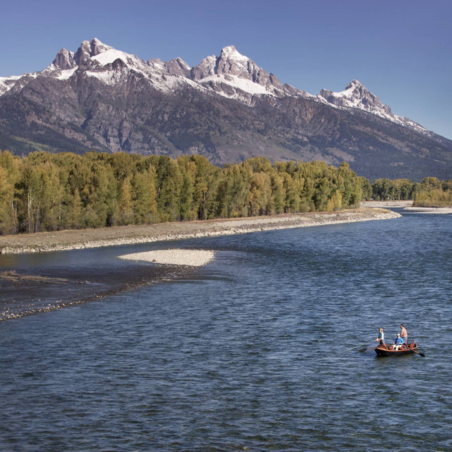 People fishing in boat on wide river by rocky shore, green trees, mountains
