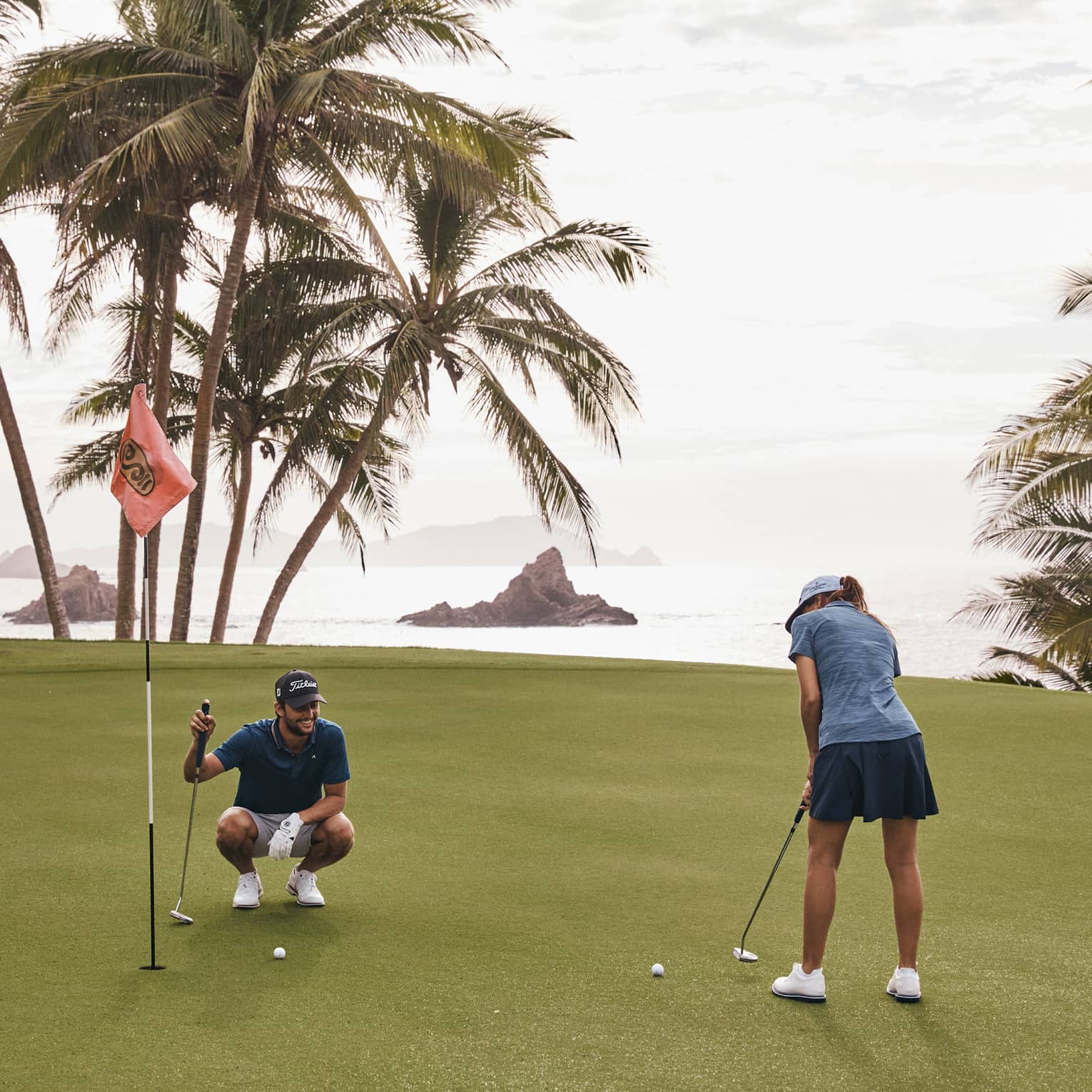 On a golf green surrounded by water and palm trees, one golfer putts while another kneels beside the hole and watches.