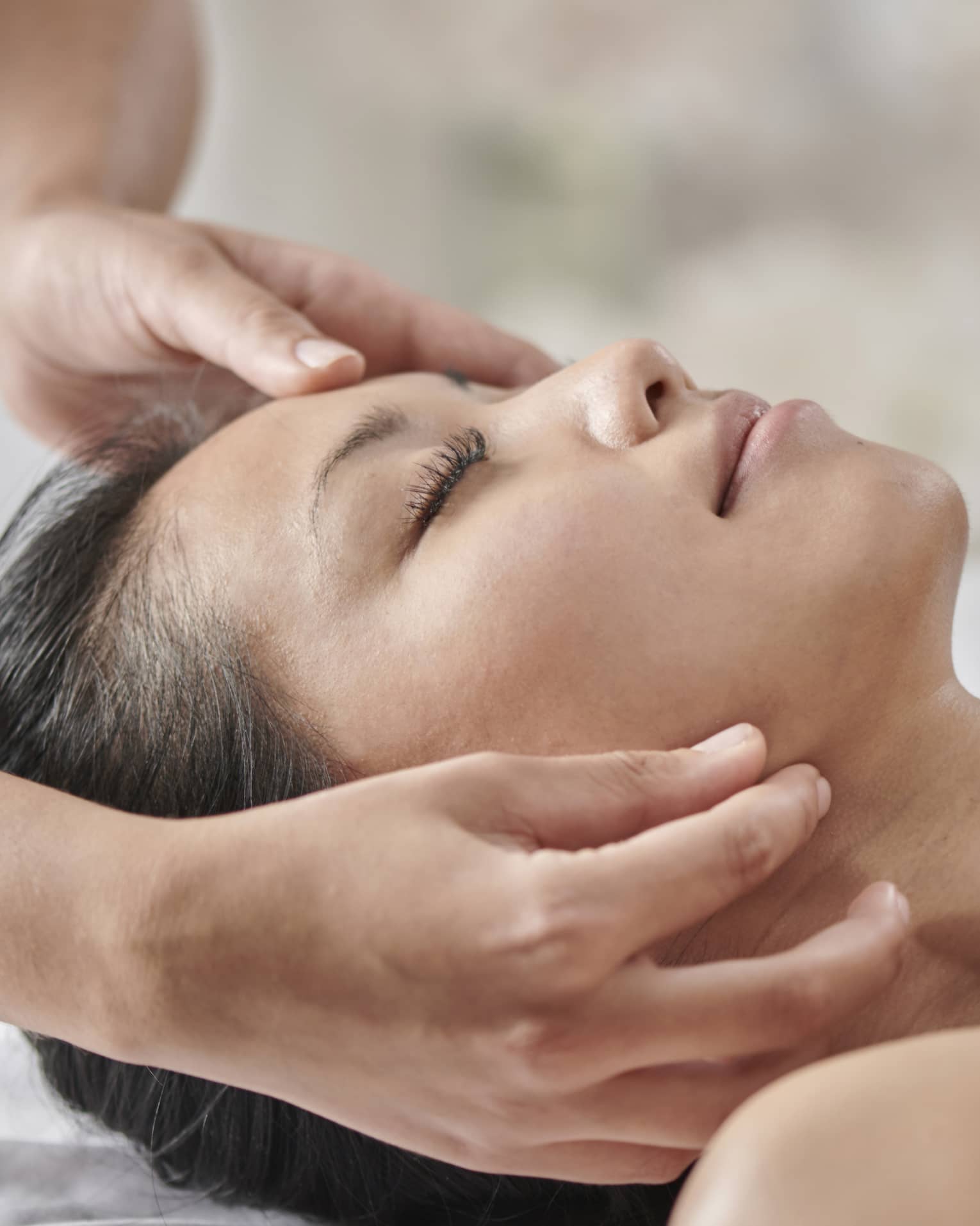 Masseuse massages woman's face as she lies face up on table under white sheet