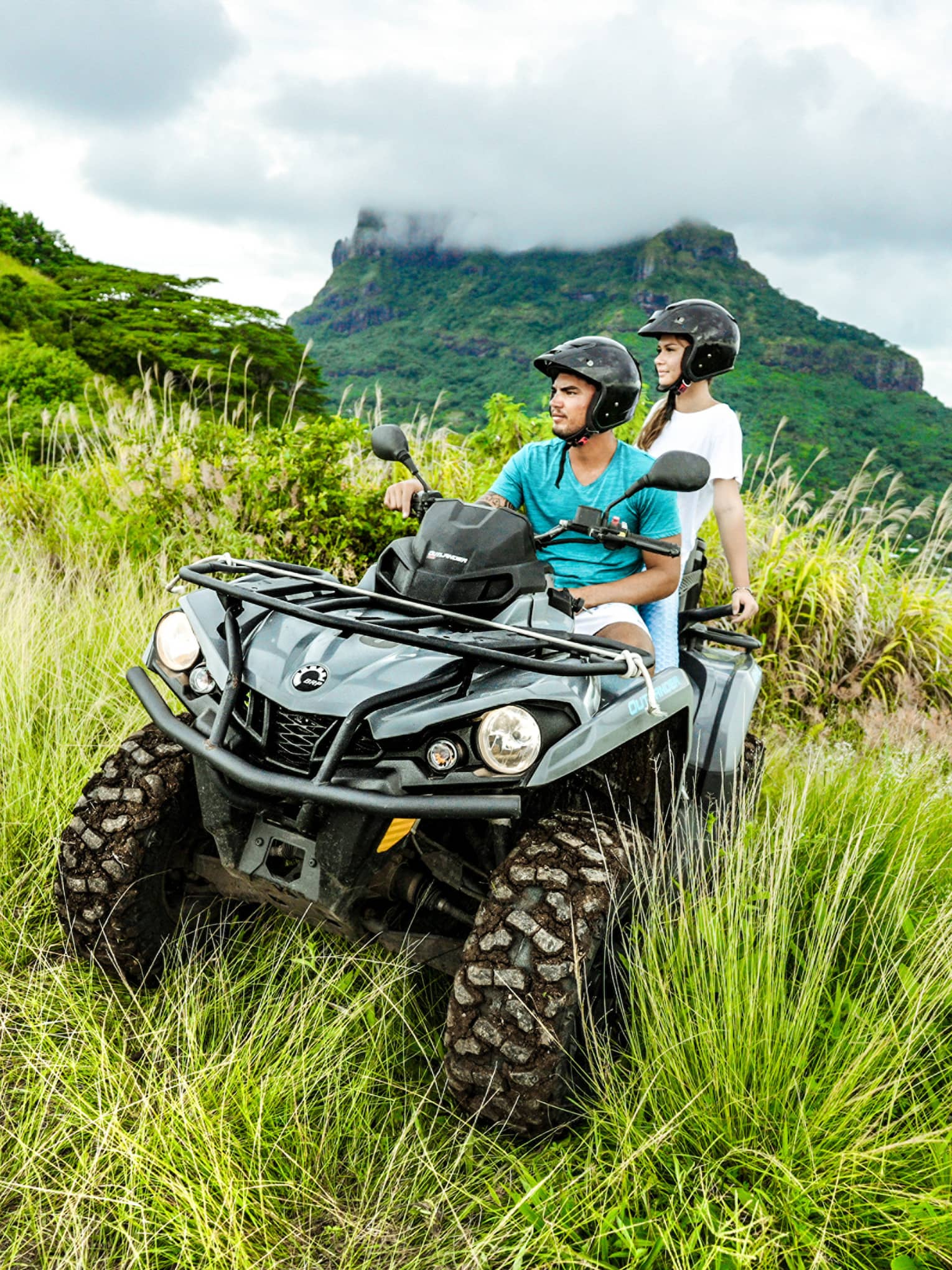 Close-up of a couple riding an ATV through lush vegetation; behind them, a plume of smoke billows above a volcanic mountain.