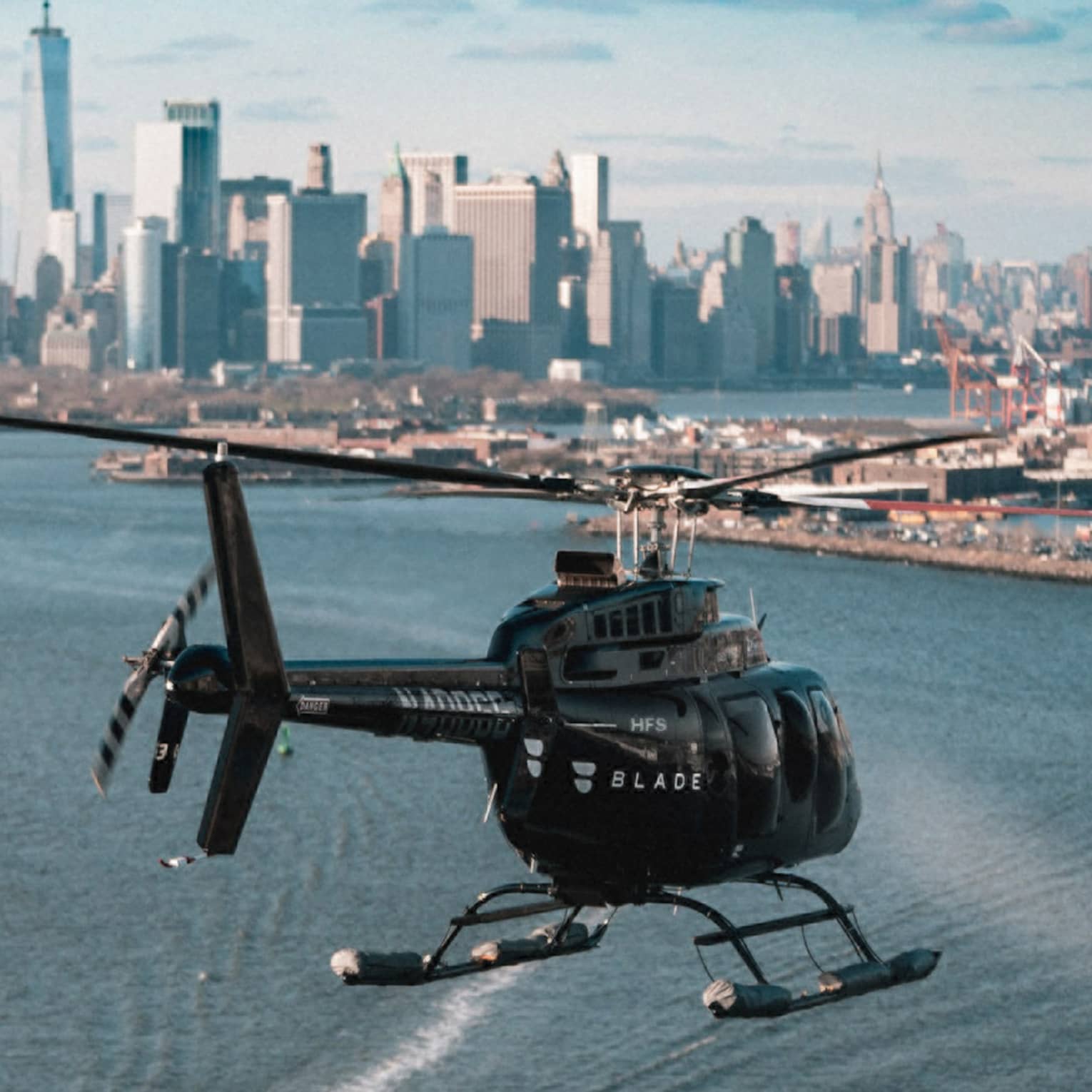 Rear view of a helicopter flying over water in the direction of a shipyard and the iconic New York skyline in the distance.