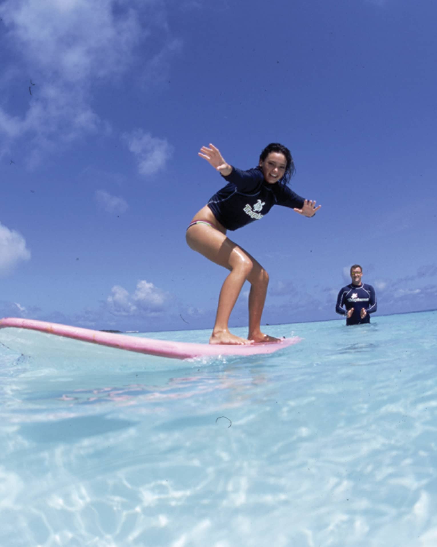 In the crystal-clear shimmering water is a woman learning to surf and an instructor standing in waist-deep water clapping.