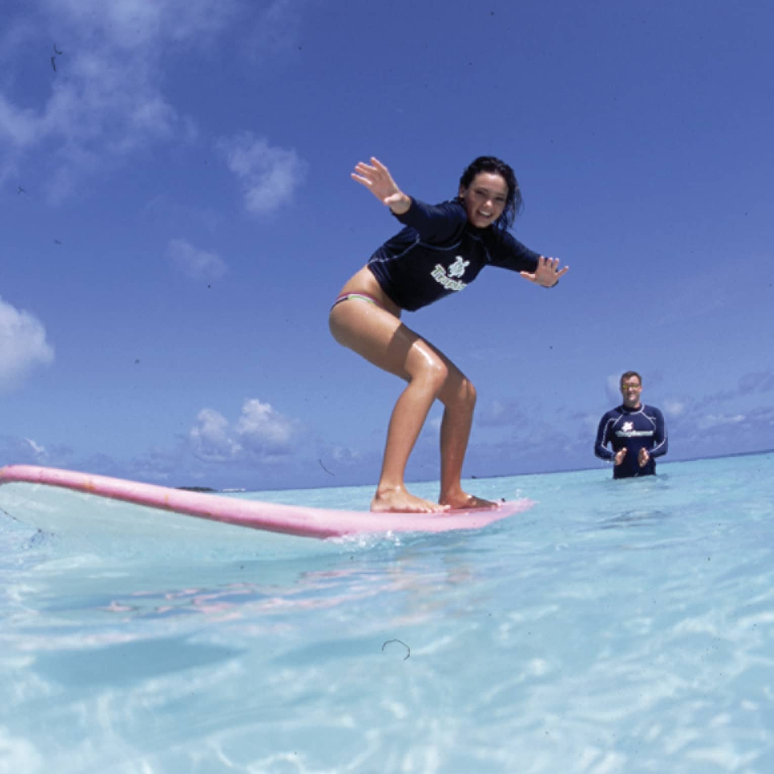 In the crystal-clear shimmering water is a woman learning to surf and an instructor standing in waist-deep water clapping.