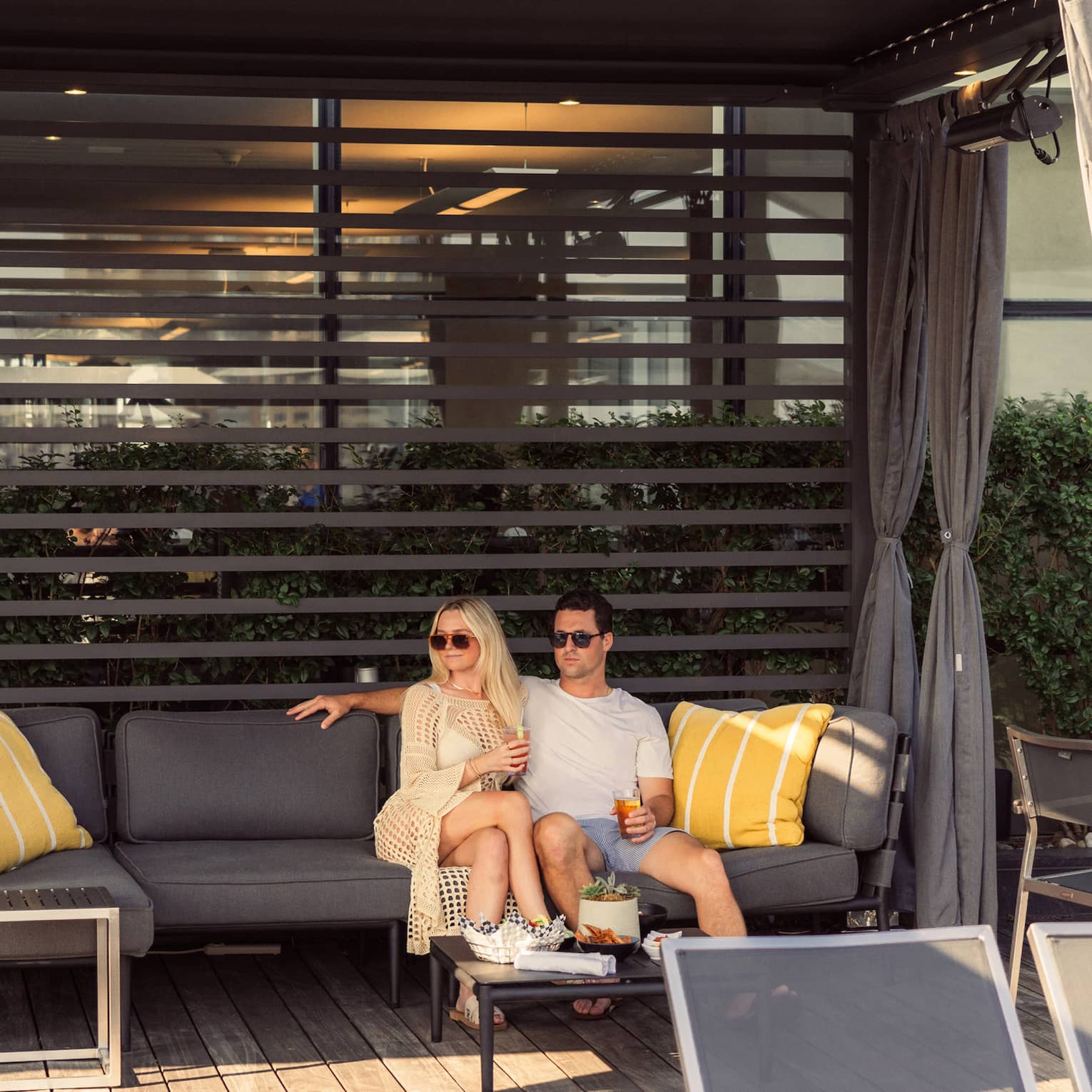 In a sun-dappled cabana, a couple relaxes together, drinks in hand, on a plush patio sofa, a table of snacks set for them.