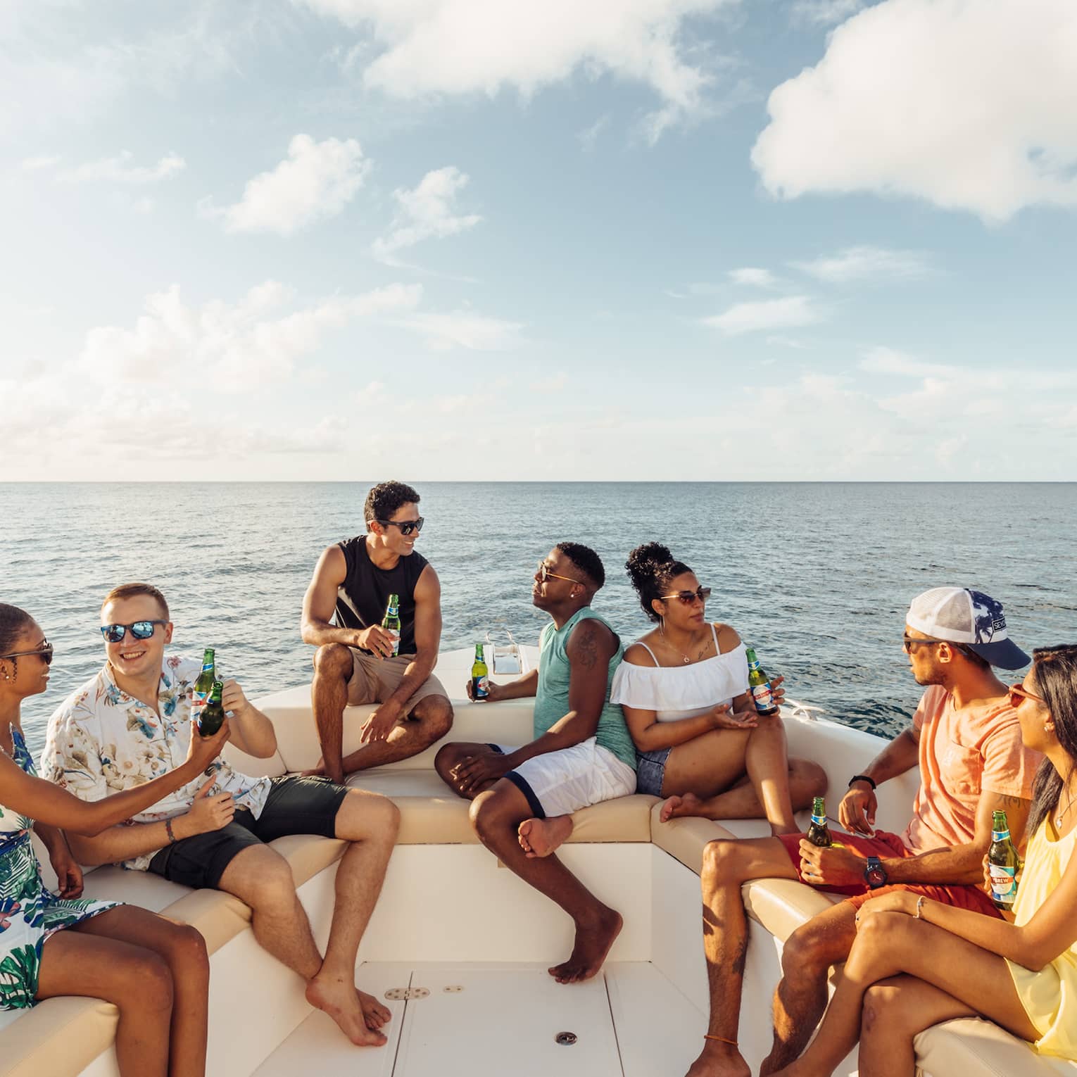 Group of guests lounge, drinks in hand, on couches lining the stern of a luxurious boat, framed by blue sky and open water. 