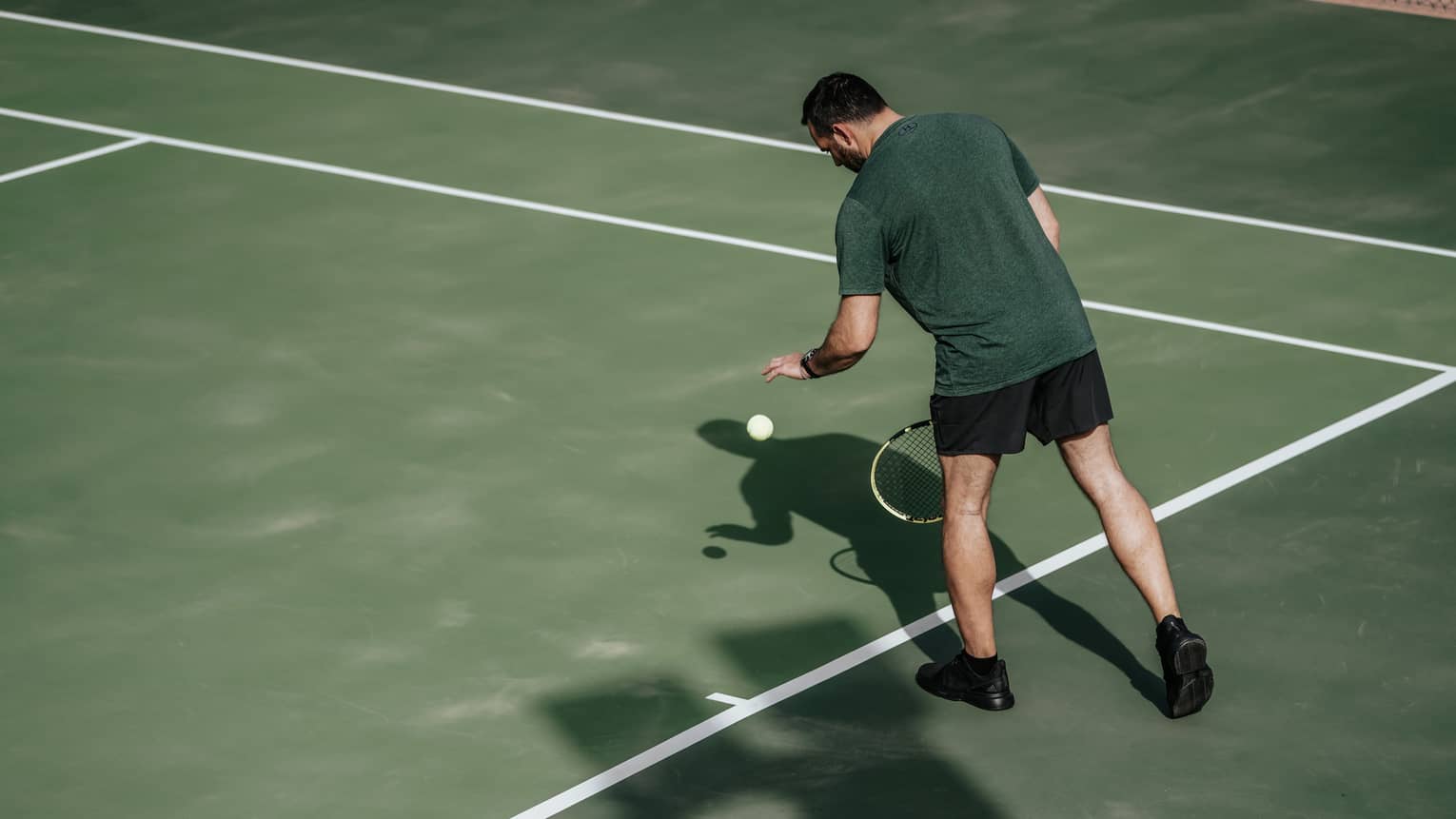 Man on tennis court bouncing tennis ball with left hand, preparing to serve