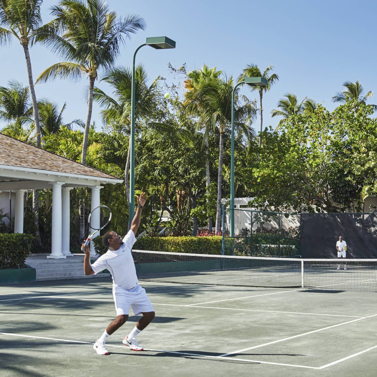A man in all white trows a tennis ball high into the air, mid-serve 