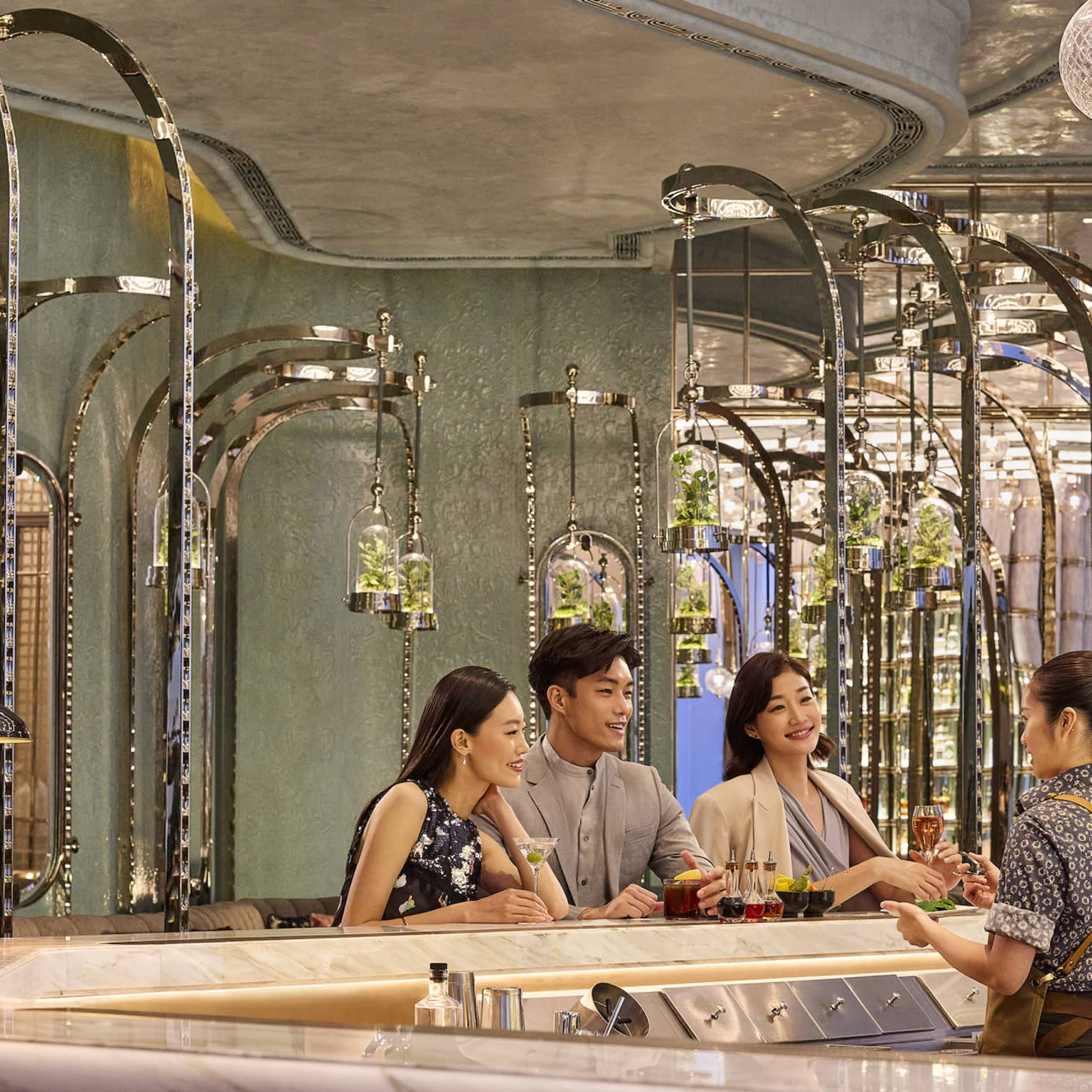 A bartender chatting with guests at the bar, each with a different cocktail, and terrariums hanging behind them on poles.