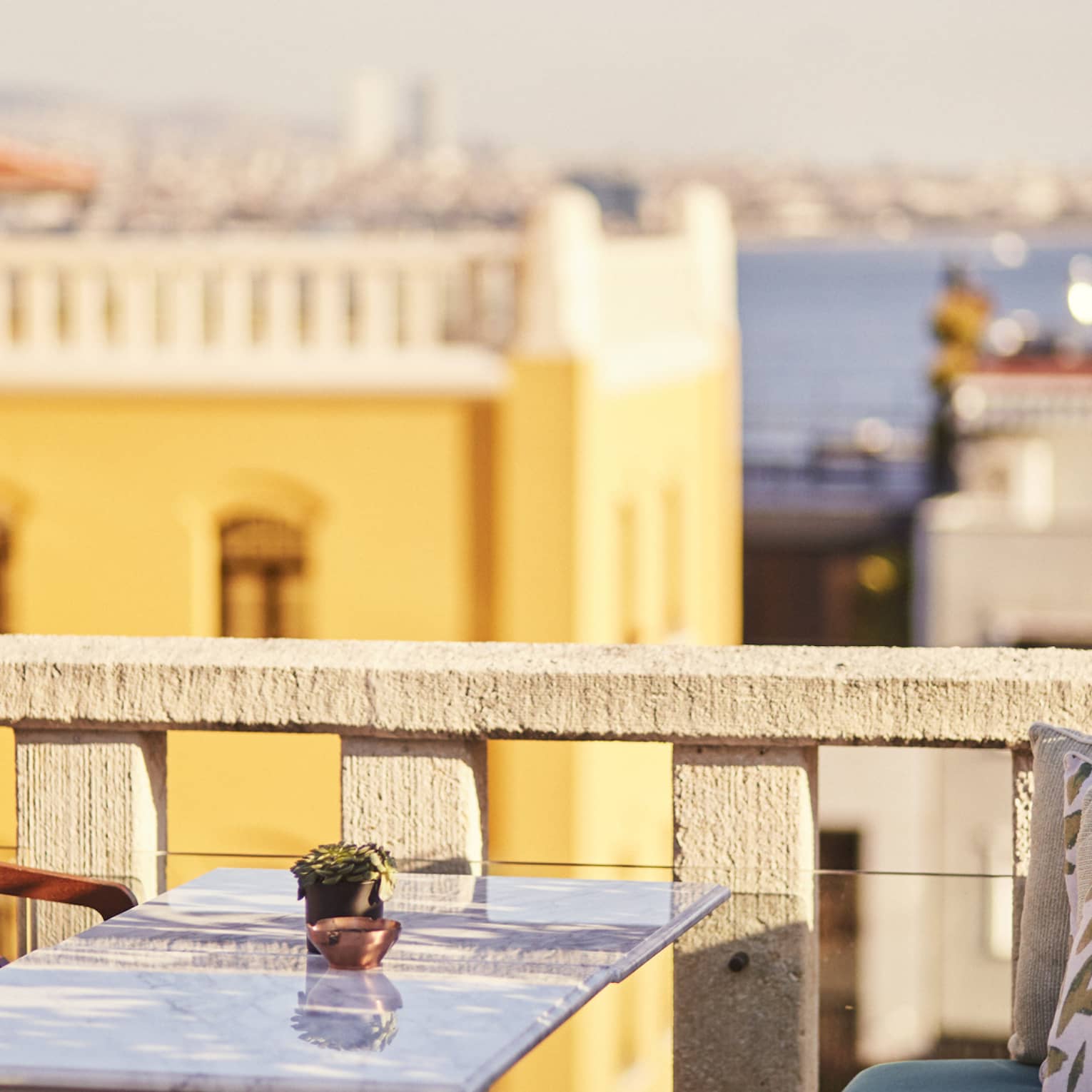 Marble table flanked by wood chairs and bench on a rooftop patio, views of beige and yellow buildings, the sea beyond. 