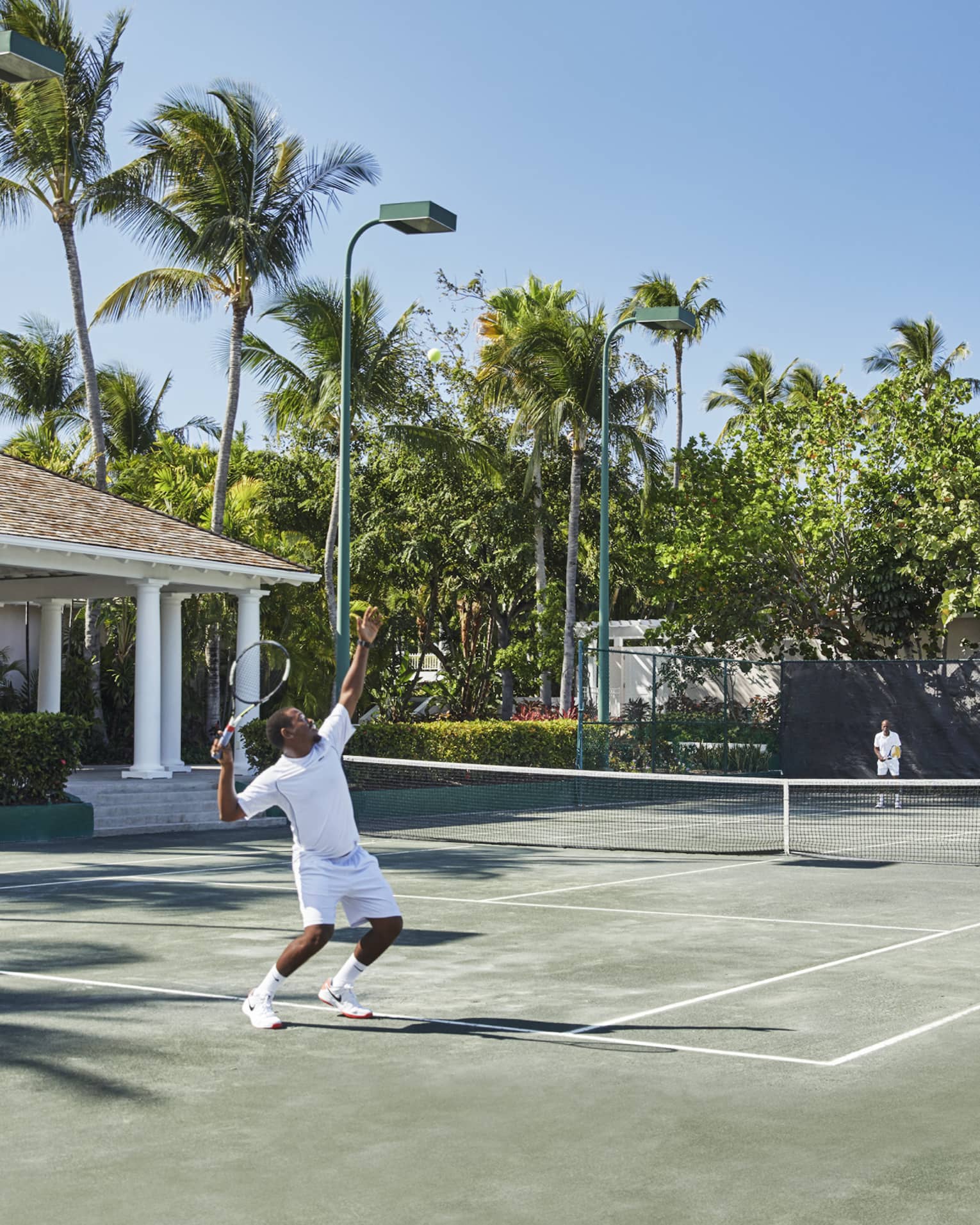 A man in all white trows a tennis ball high into the air, mid-serve 