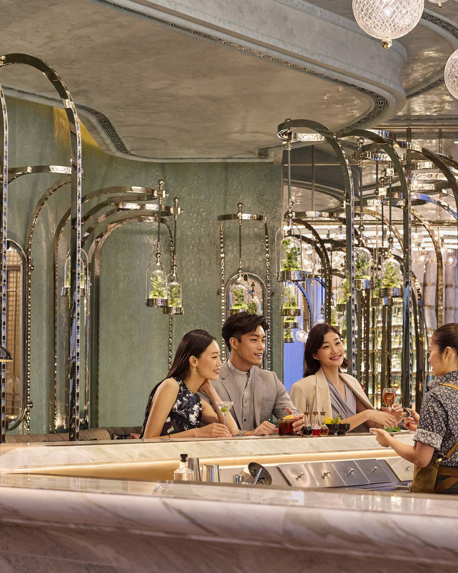 A bartender chatting with guests at the bar, each with a different cocktail, and terrariums hanging behind them on poles.