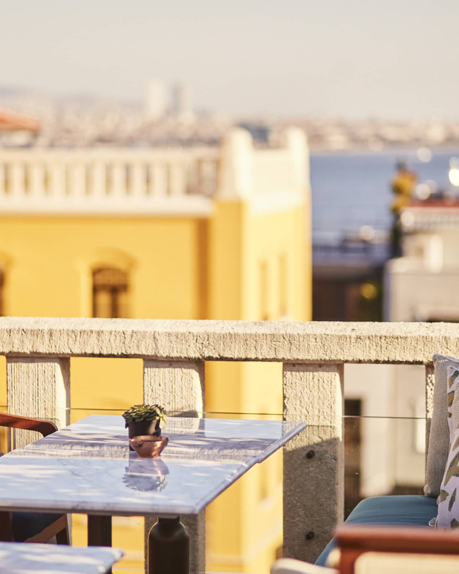Marble table flanked by wood chairs and bench on a rooftop patio, views of beige and yellow buildings, the sea beyond. 