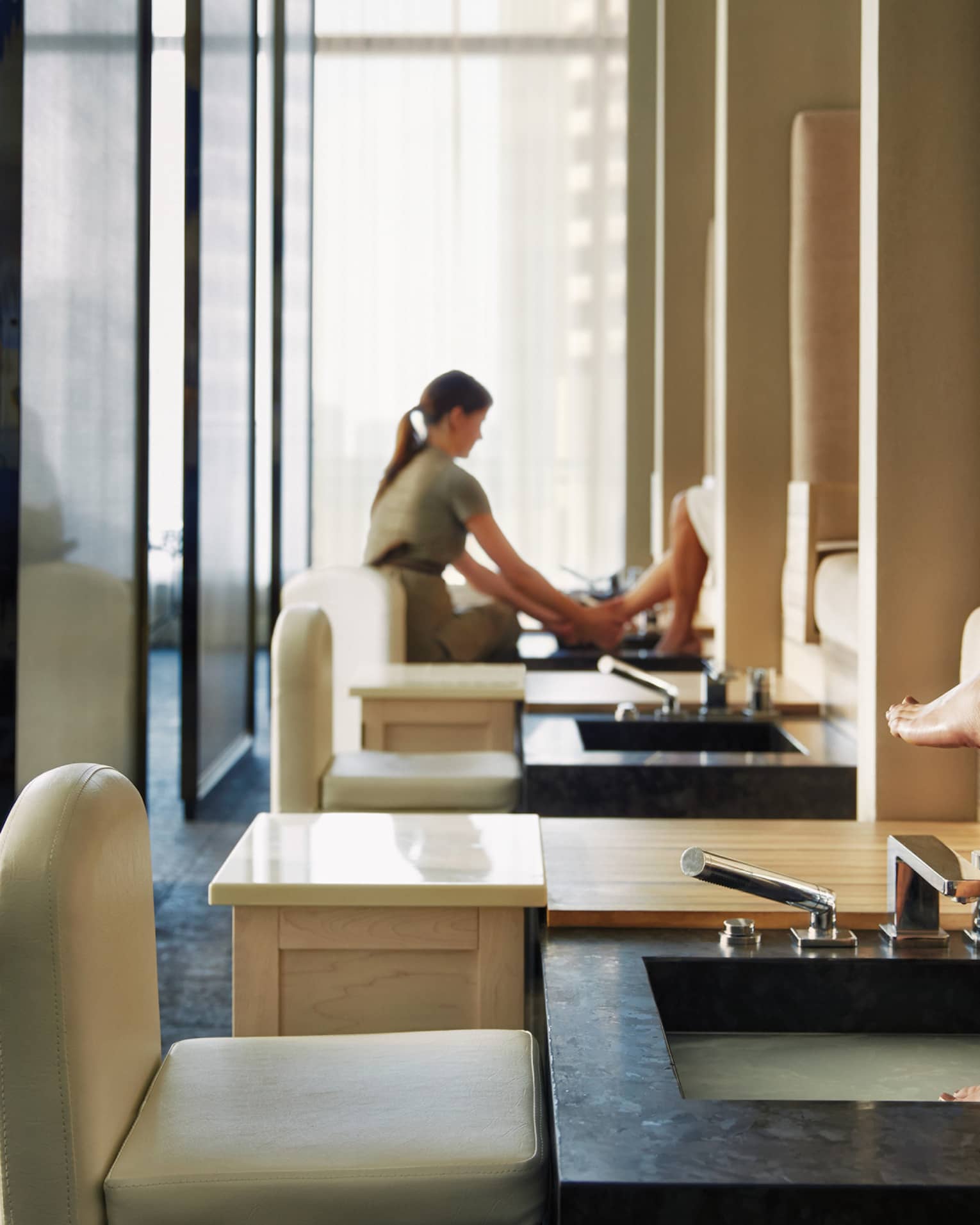 Woman's feet on pedicure sink along row of spa chairs, salon staff in background