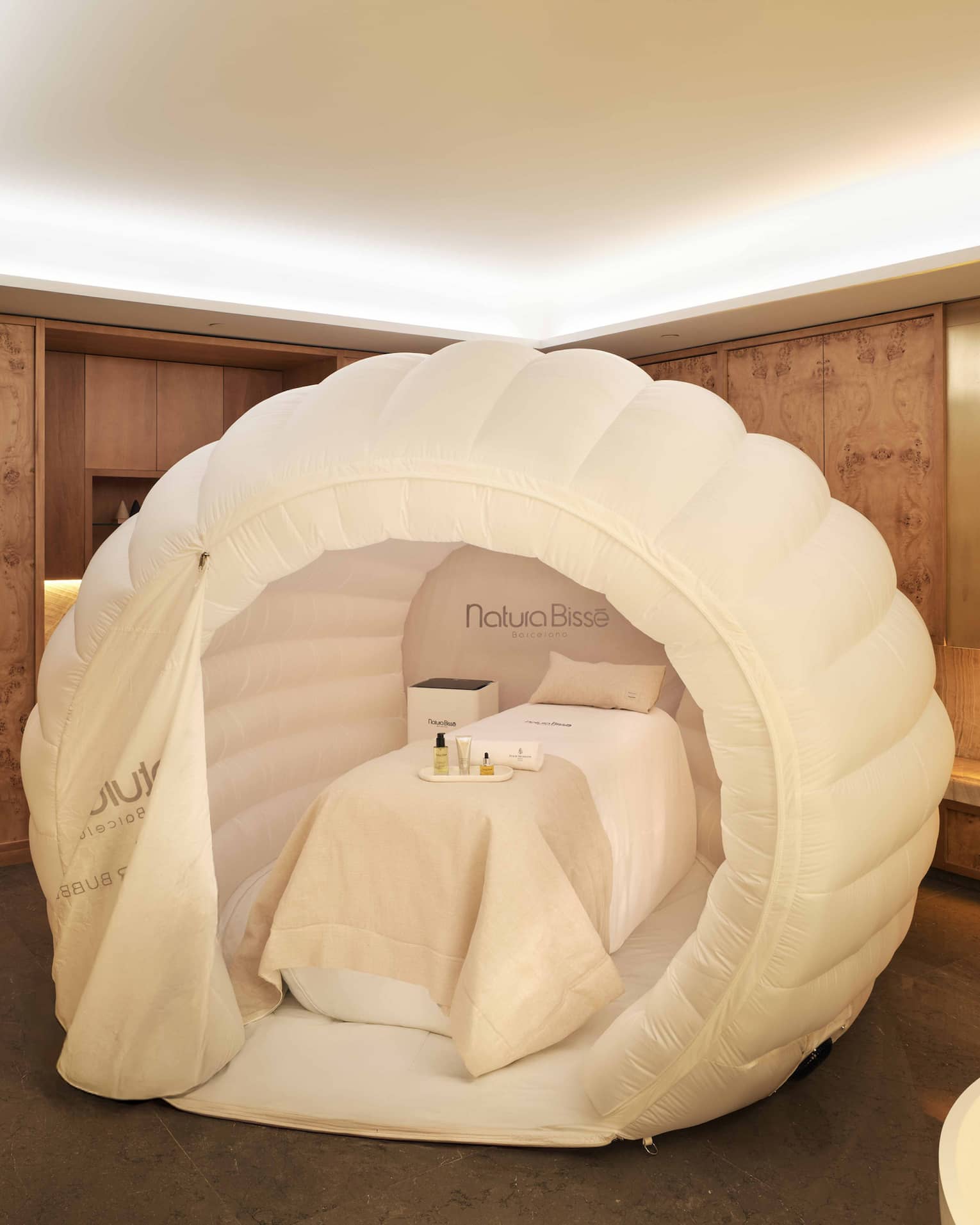 A spa treatment table with white draping and pillow within a near-spherical white tent, all in a spacious, softly lit treatment room with decor in warm earthy tones