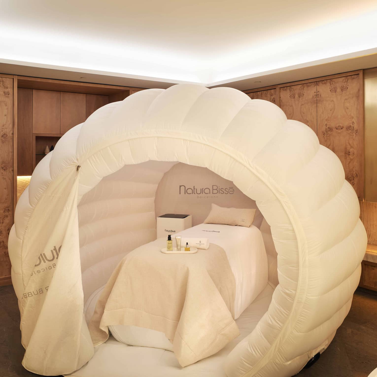 A spa treatment table with white draping and pillow within a near-spherical white tent, all in a spacious, softly lit treatment room with decor in warm earthy tones
