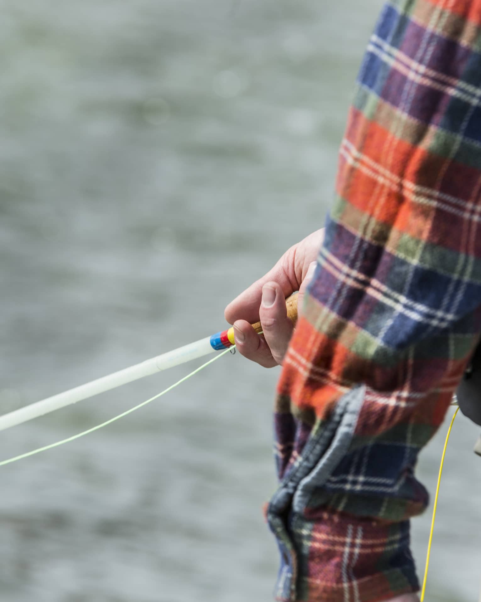 Against a background of rippling silver water, a plaid sleeve partly reveals a hand gently holding a colourful fishing rod.