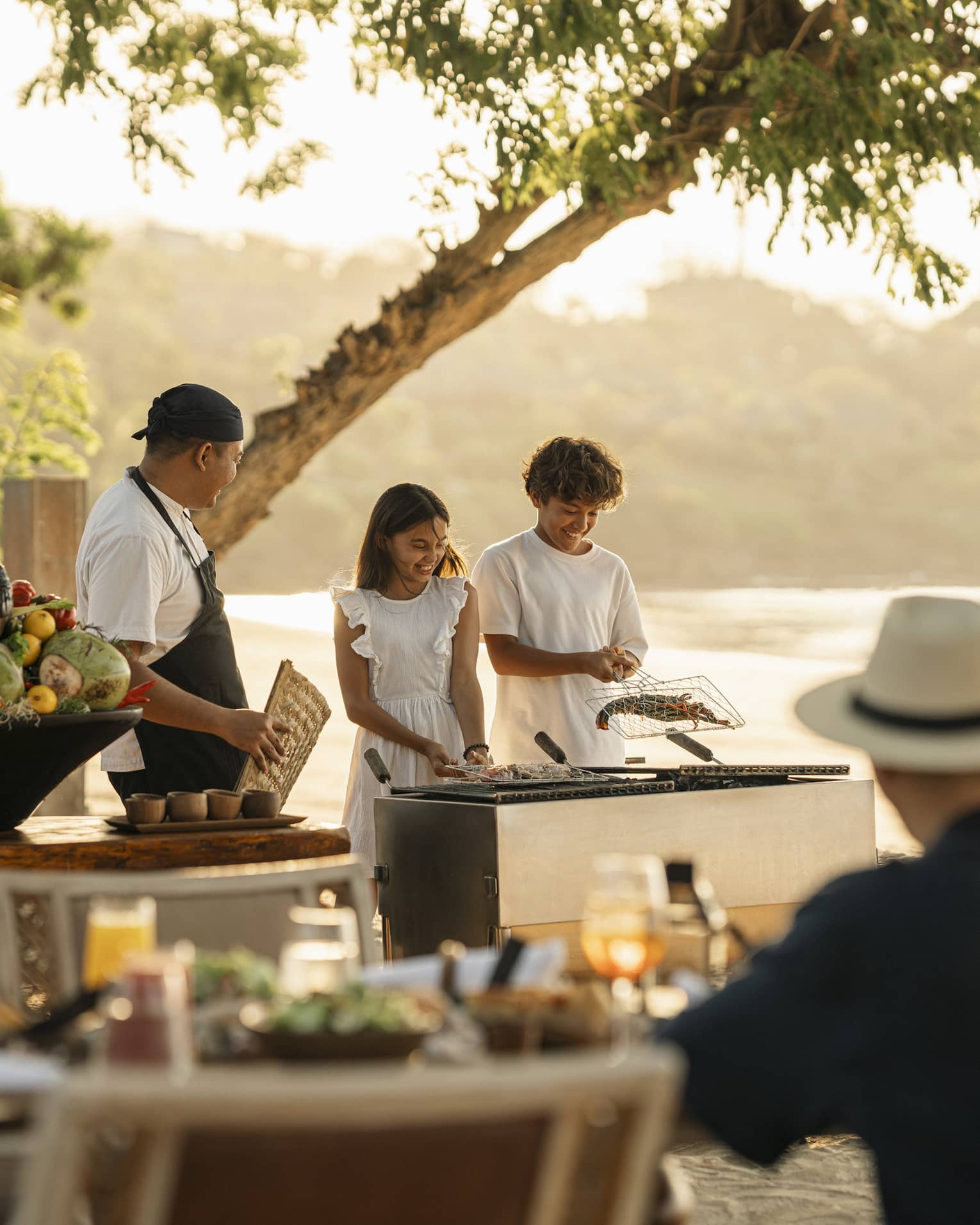 A chef smiles at two children who barbecue shellfish in a grilling basket on the beach as their parents watch.