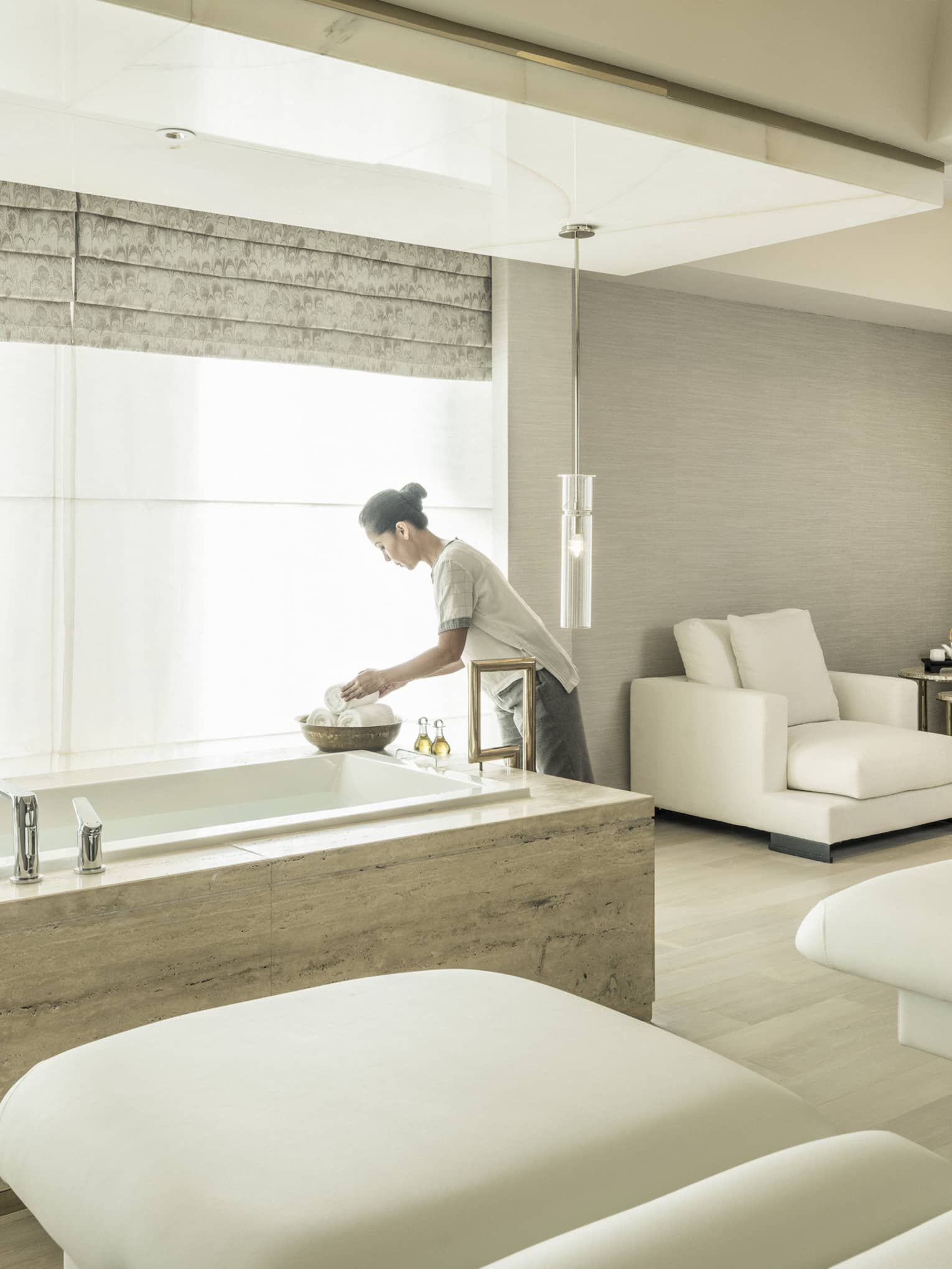 A spa attendant arranges some products in the spa suite, with two massage beds and a soaking tub in front