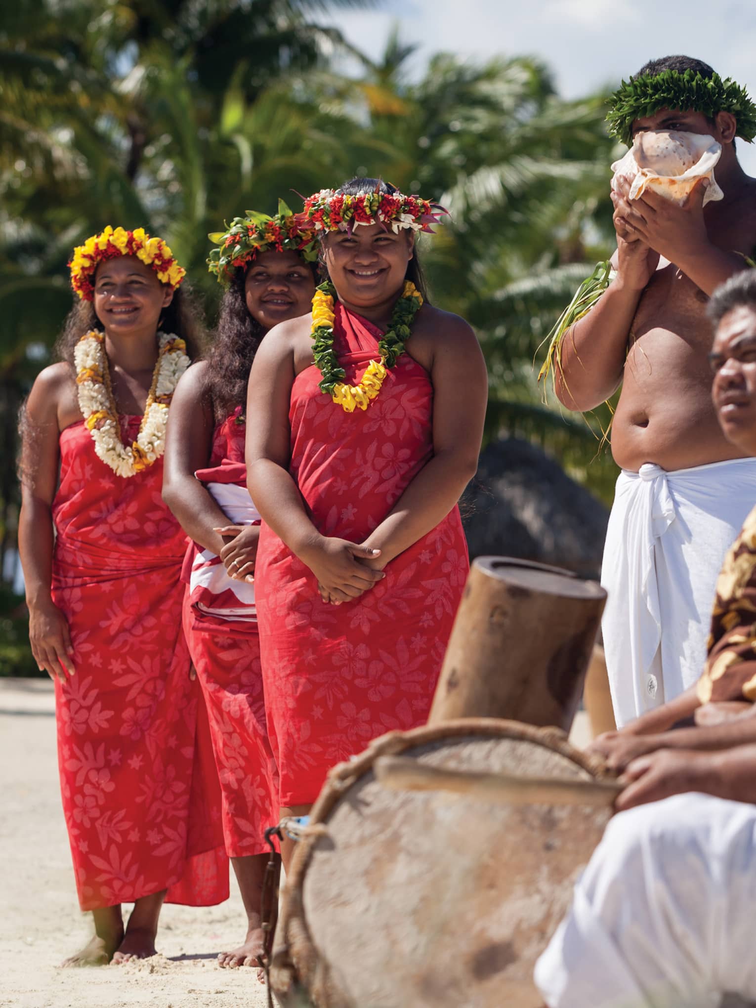 Group of people in traditional attire with leis and a conch shell, standing on a beach