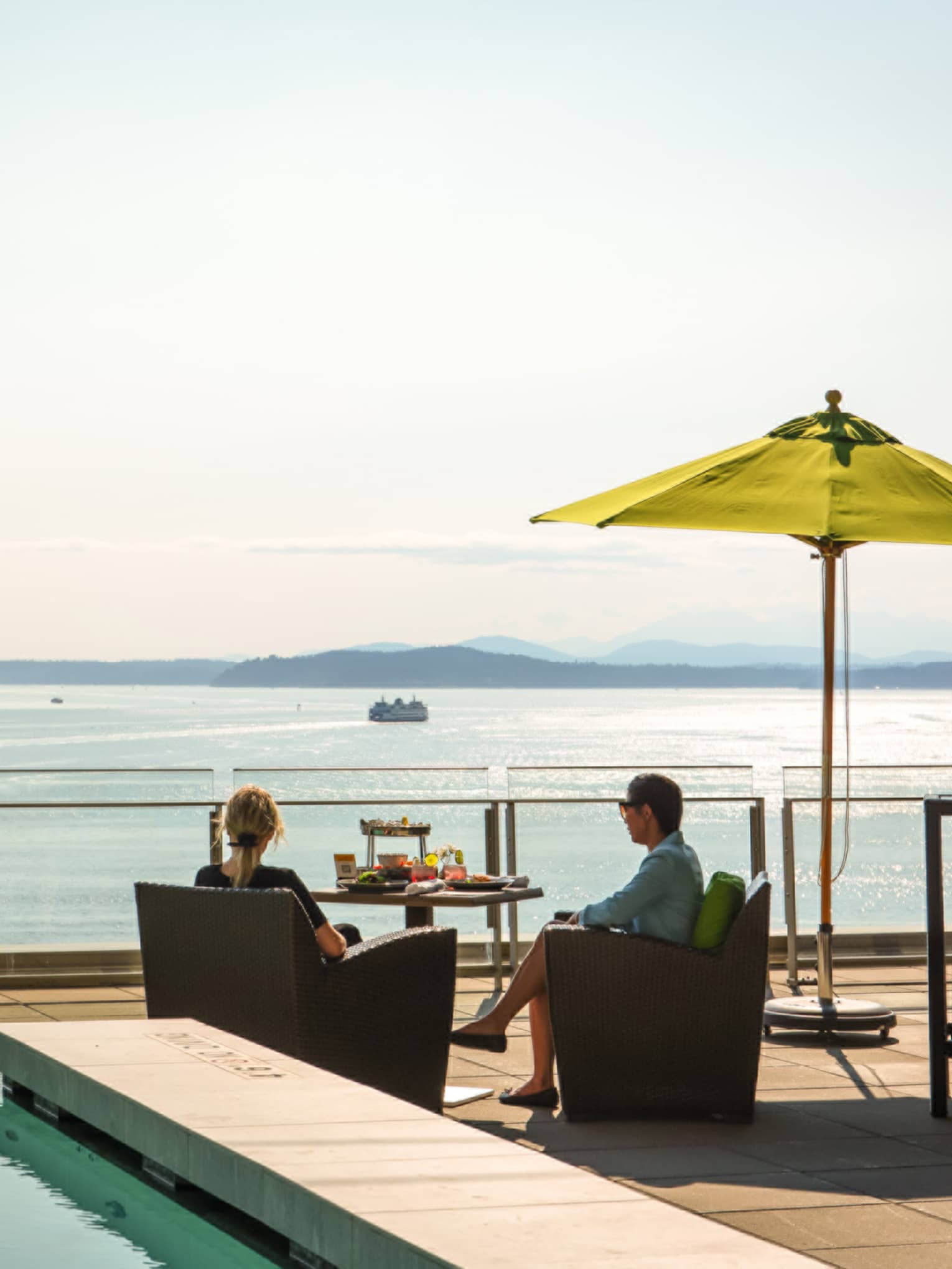 Couple sits poolside at dining table on sunny patio overlooking Elliott Bay and The Seattle Great Wheel