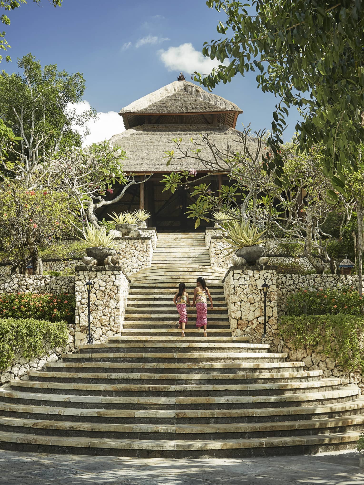 Back of two young girls walking up large stone staircase to stone lobby with thatched roof