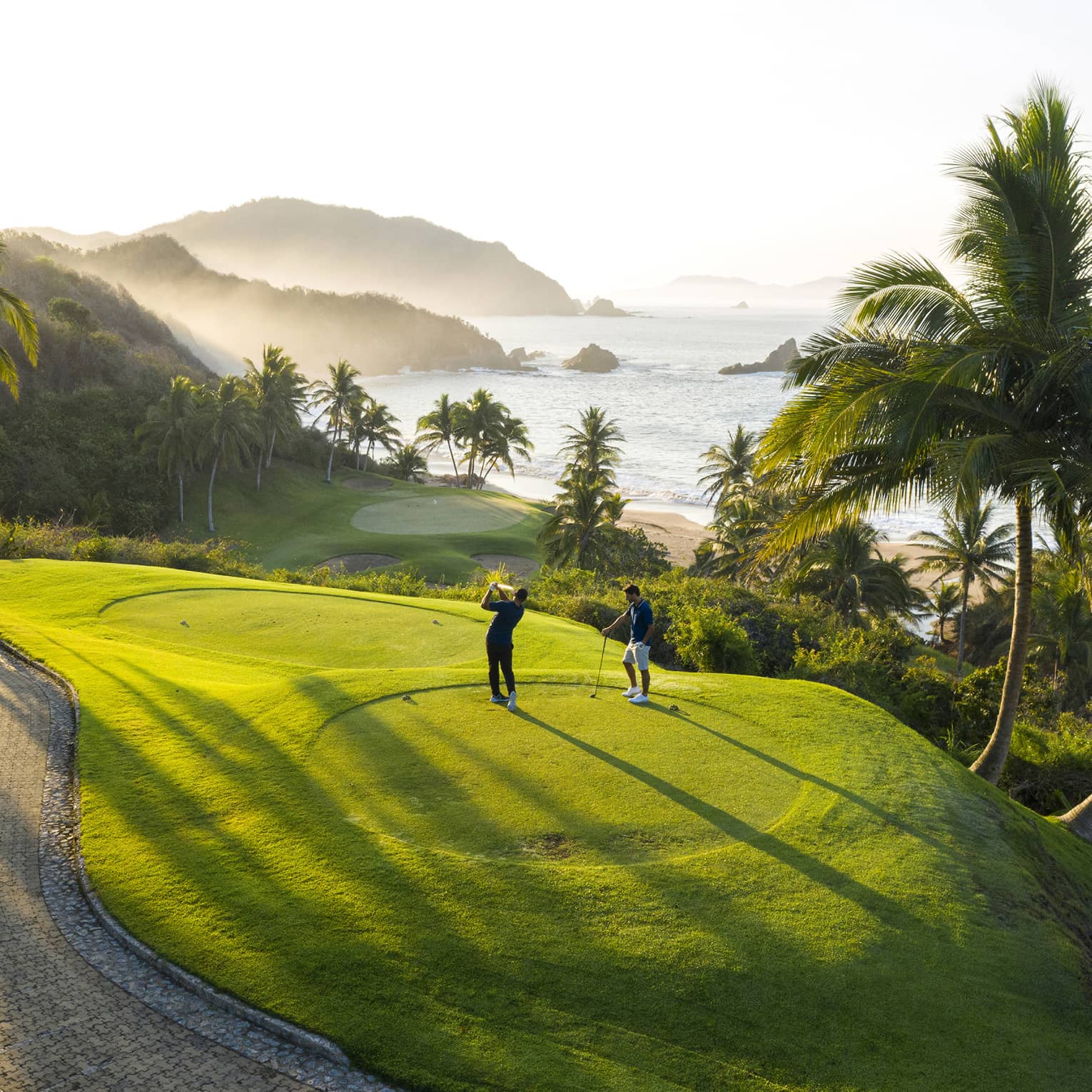Rear view of two golfers on an elevated tee, one swinging toward the green down by the beach, misty hills and ocean beyond.