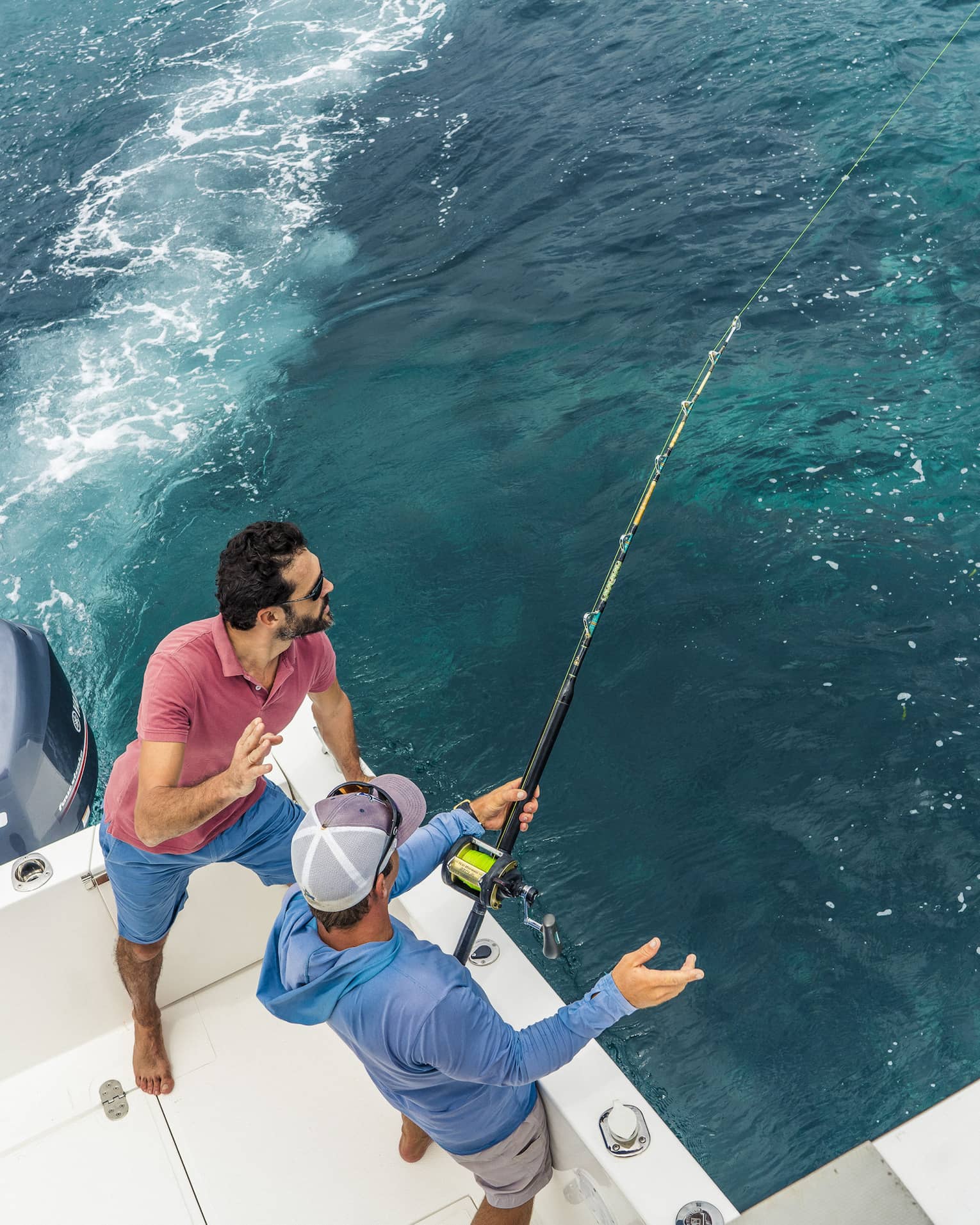 Aerial view of two people fishing off the back corner of a motorboat. One grips a fishing rod while the other looks on.