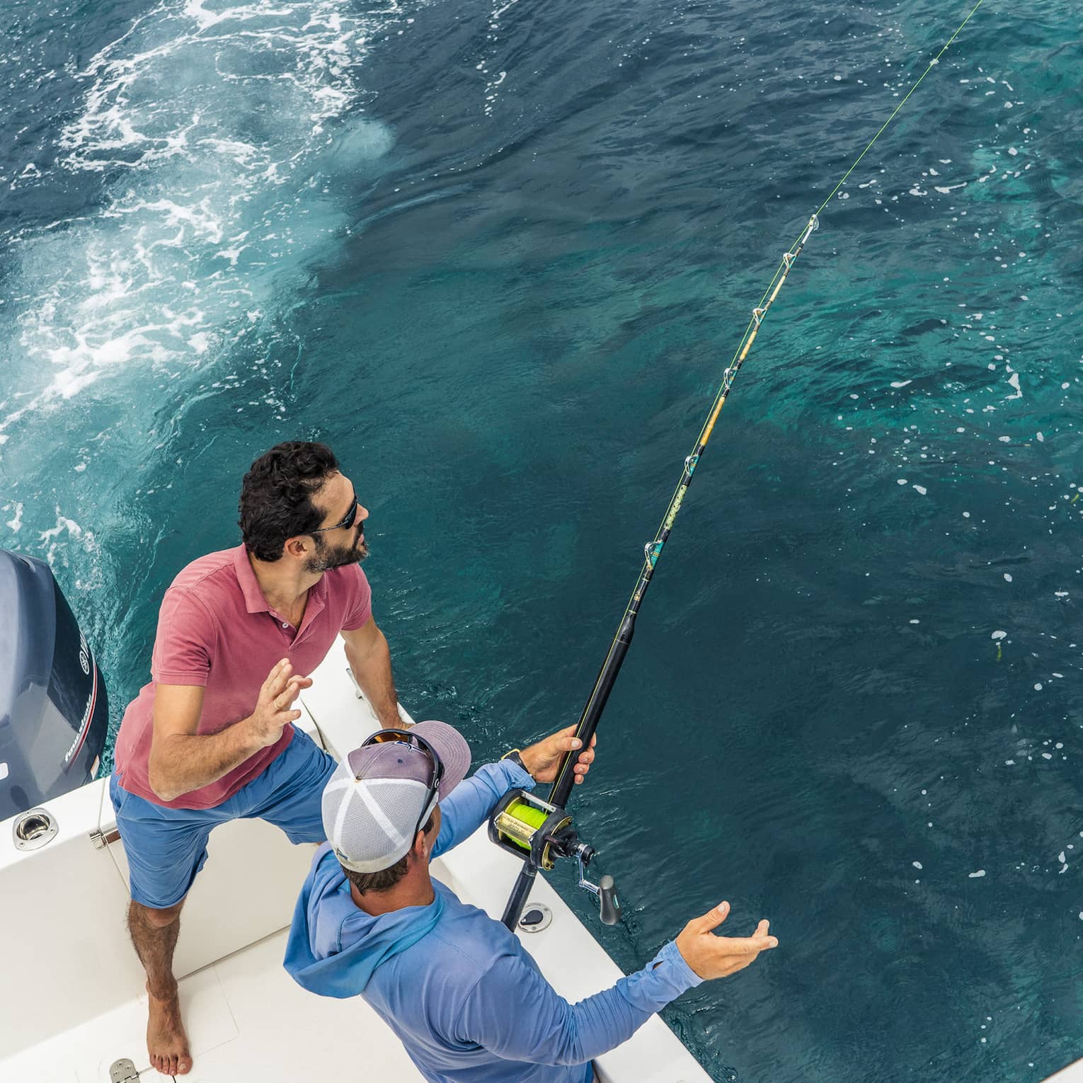 Aerial view of two people fishing off the back corner of a motorboat. One grips a fishing rod while the other looks on.