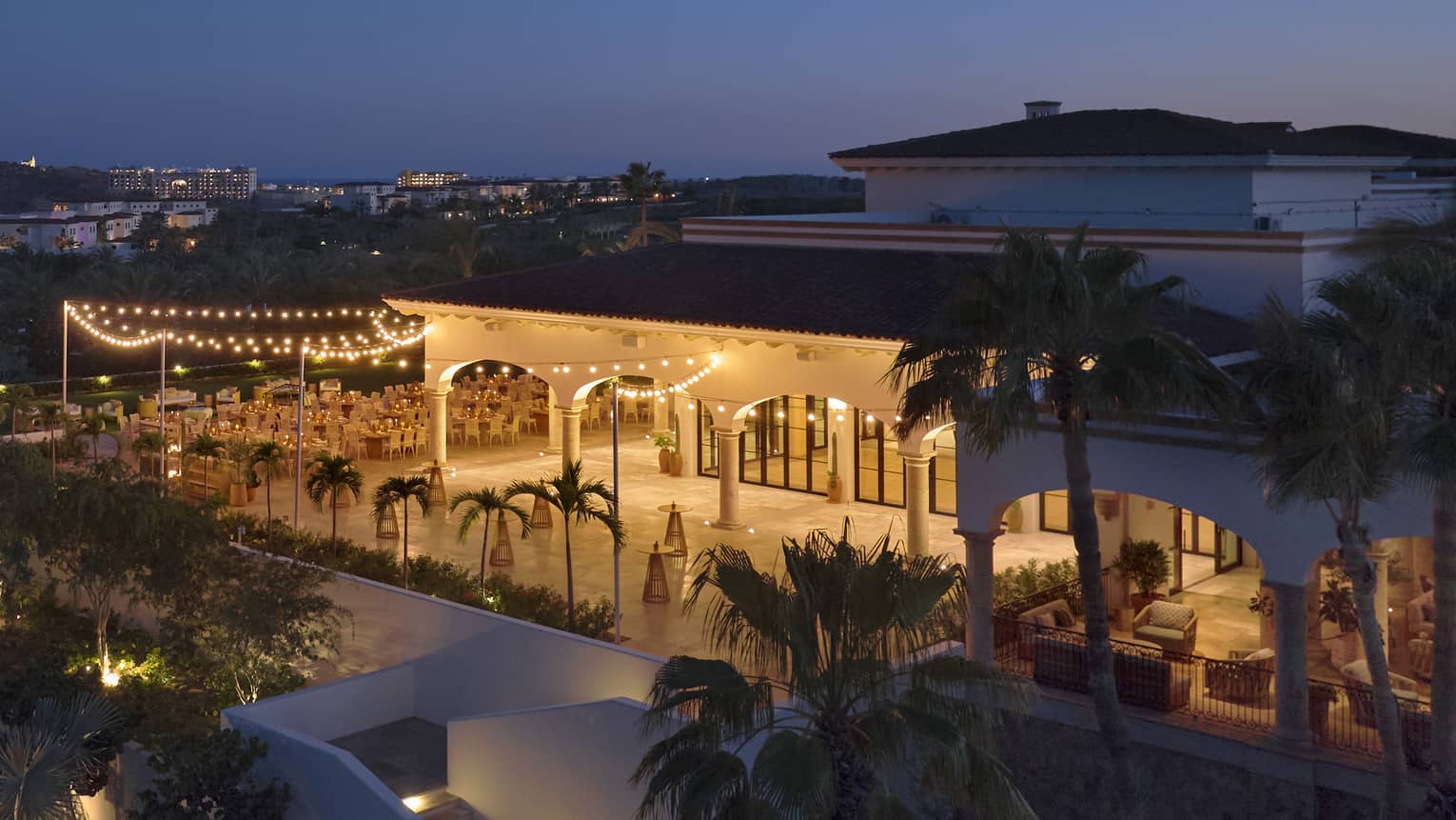 A an outdoor event space brightly lit in the evening, set among palm trees and next to a building