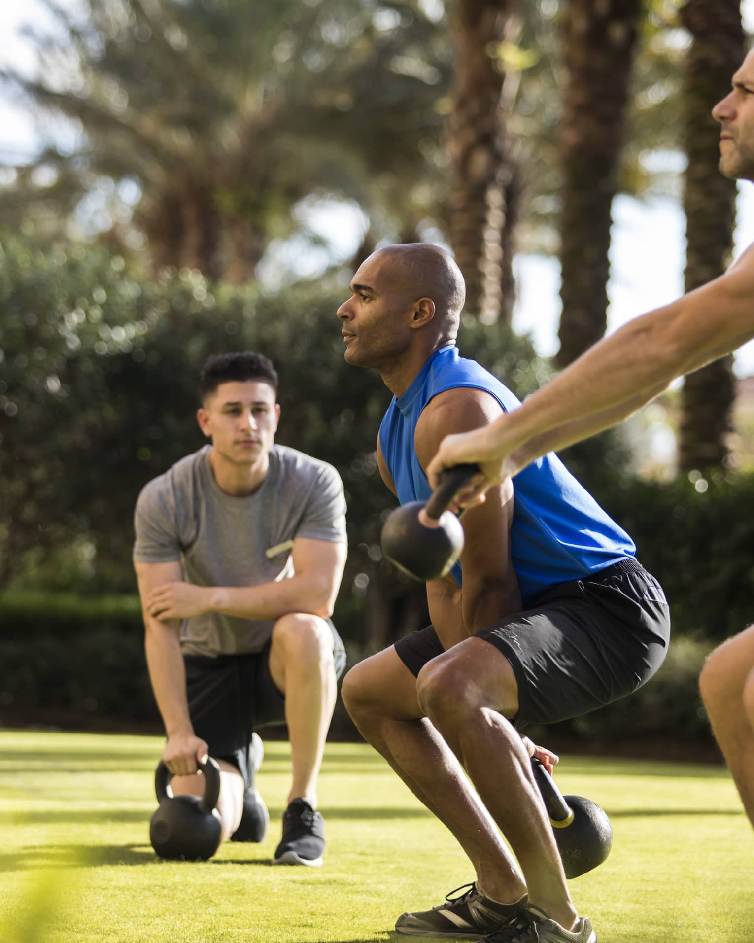 Four Seasons trainer watches as two men squat, lift kettlebell weights on lawn