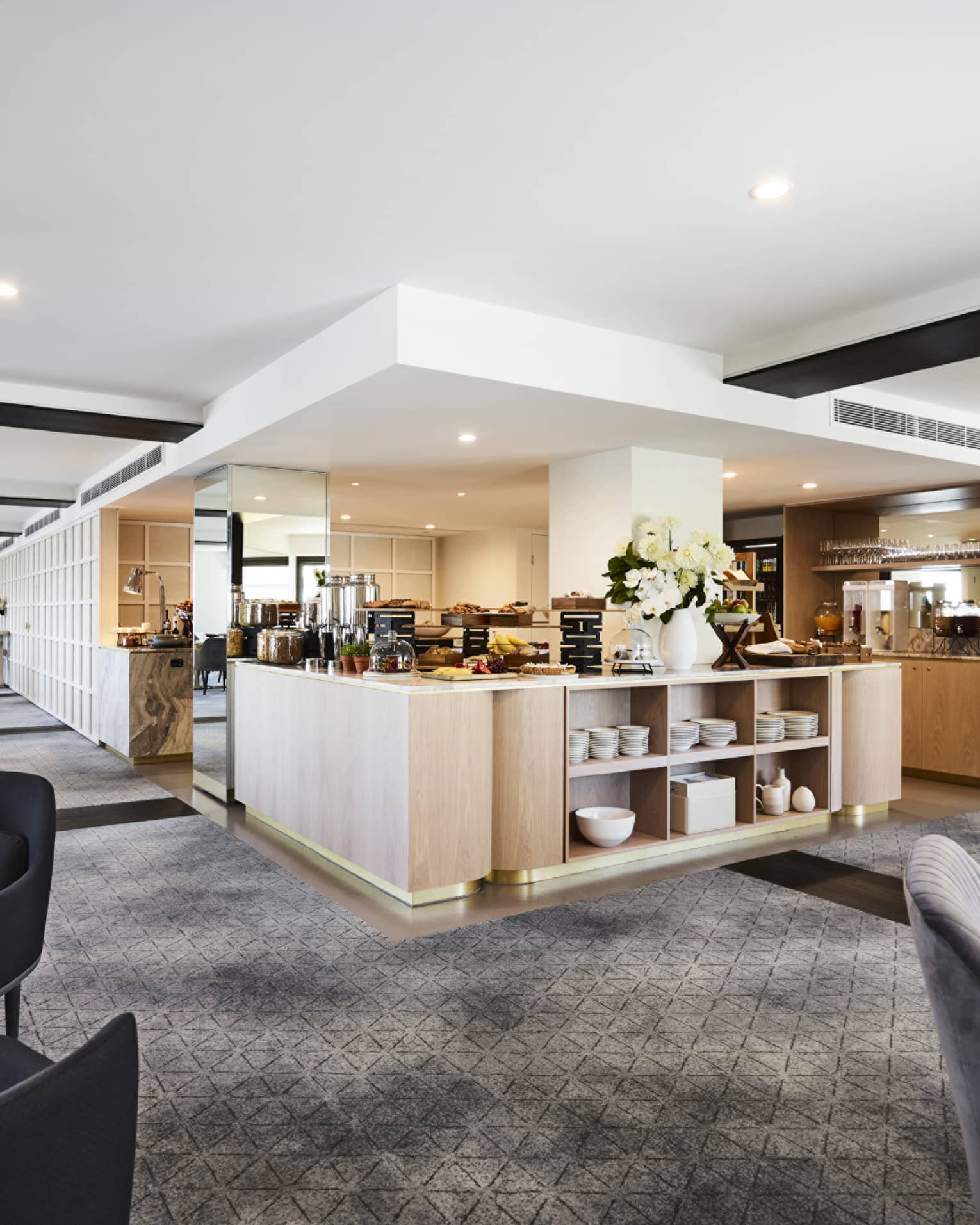 A modern hotel lounge with plush grey chairs and carpet, and a well-equipped kitchen set up with buffet foods and coffees.