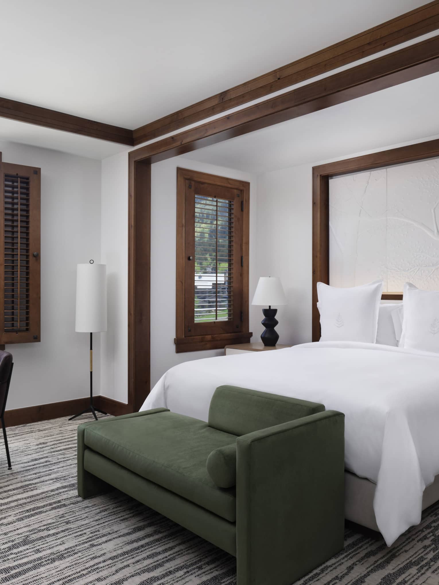 Resort room with a king bed, green chaise, nightstands with lamps, dark wood accents, a table and chair by the window with mountain view