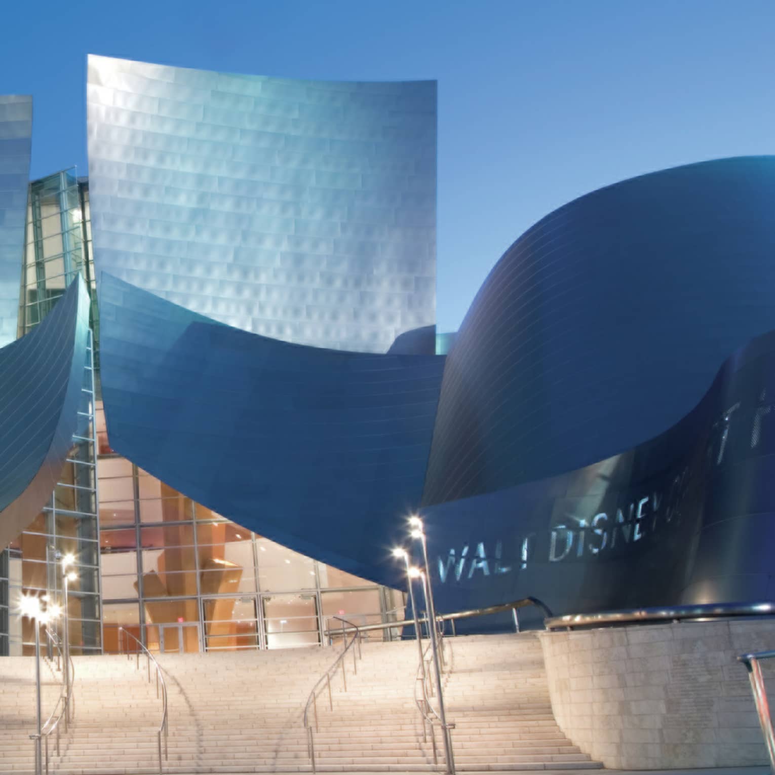 Enormous, curved metal wall and roof structures shine in the light emitted by lamps on the front steps of a concert hall.