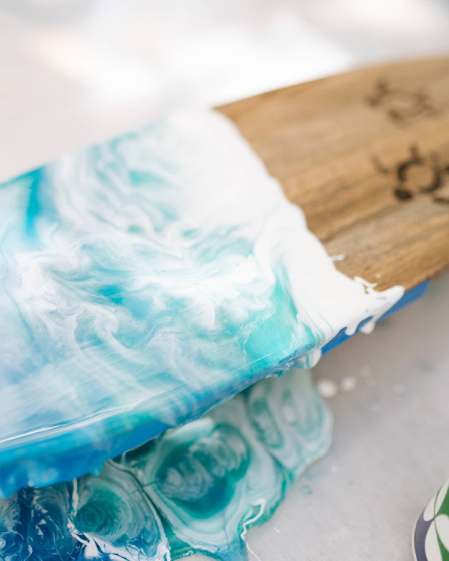 Close-up of wooden surfboard half-painted in deep to turquoise blues with white wave-like swirls and turtle outline drawings.