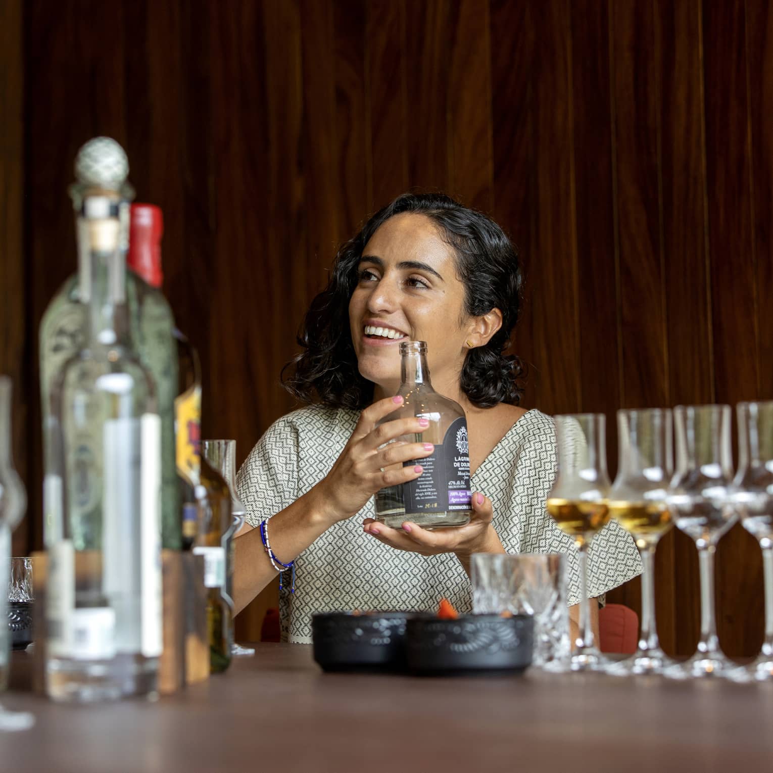 A guest cradles a short, wide bottle of liquor at a table littered with bottles of various sizes and grappa glasses. 