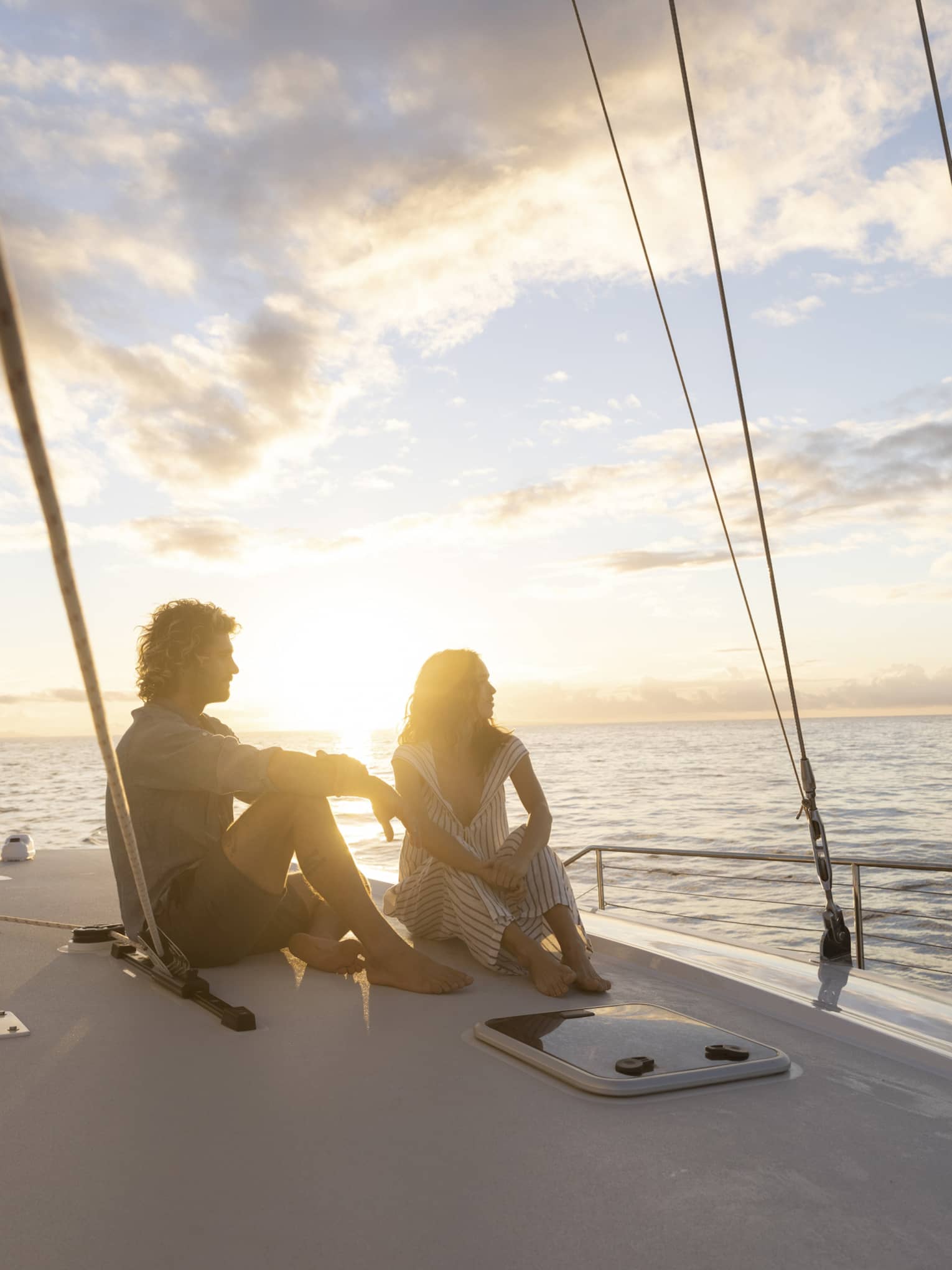 Couple in silhouette sitting barefoot on a boat deck framed by rigging ropes against a bright blue clouded sky at sunset.
